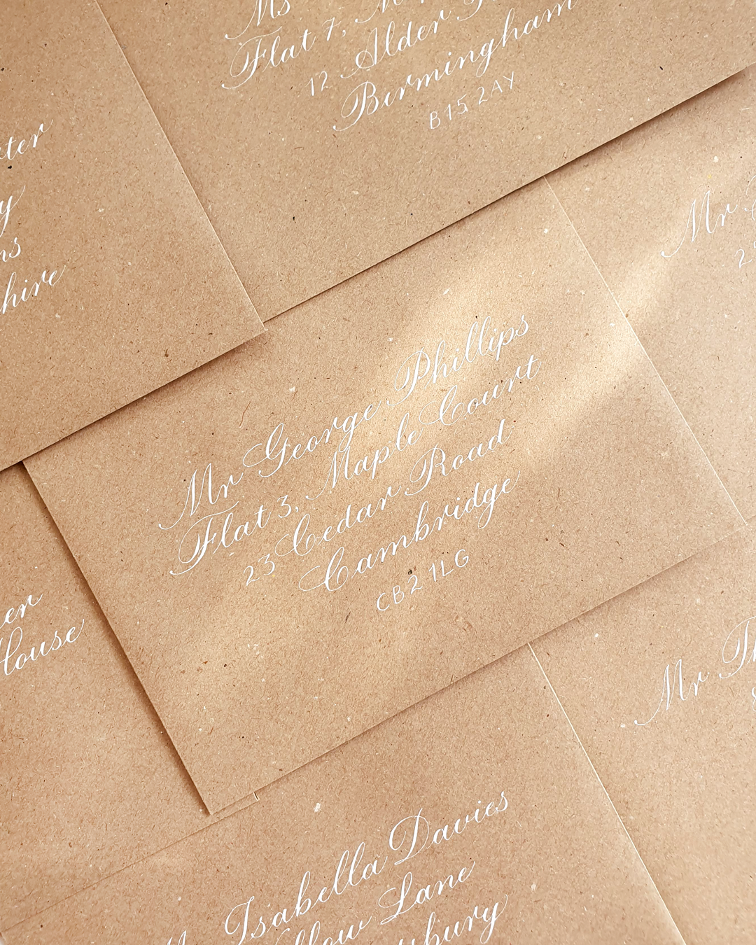 Kraft brown envelopes are shown in close up with addresses written on the front of each in copperplate calligraphy with white ink.