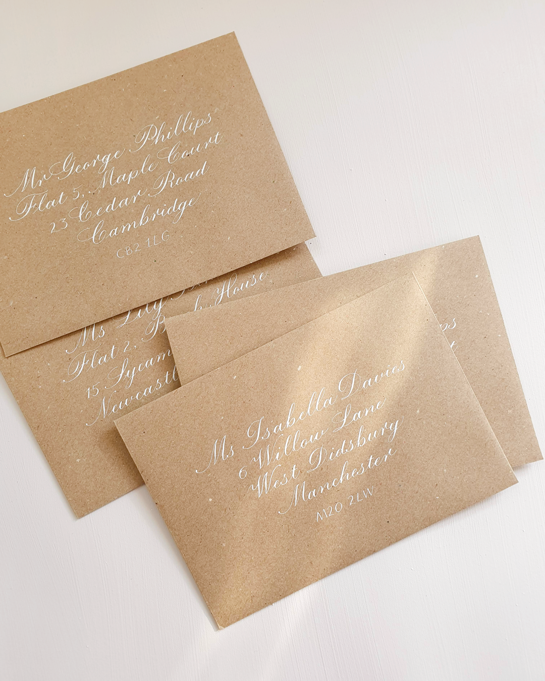 Four kraft brown envelopes rest on white background with addresses written on the front of each in copperplate calligraphy with white ink.