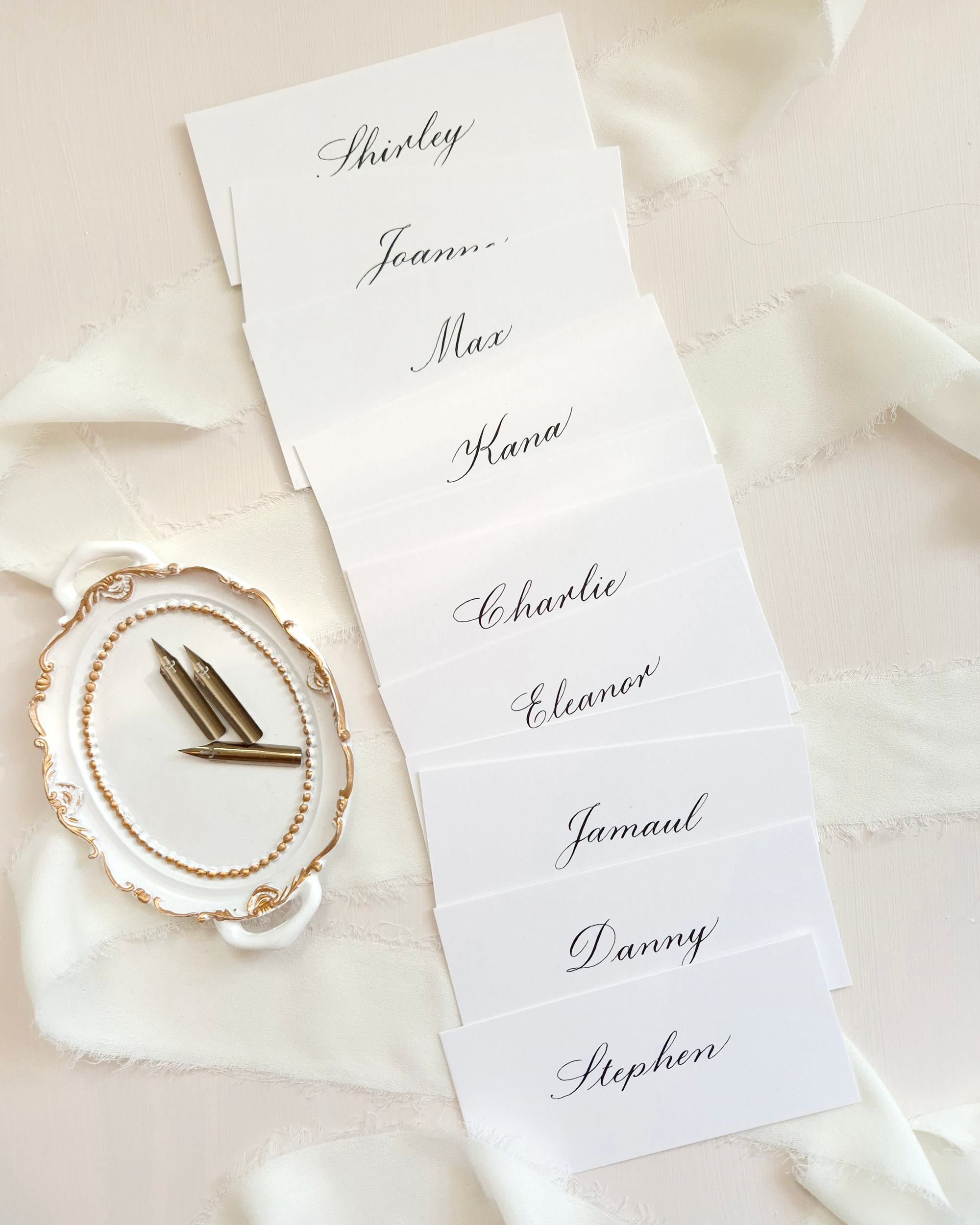 A stack of white place cards with black calligraphy names written on them. The names are: Shirley, Joan, Max, Kana, Charlie, Eleanor, Jamaul, Danny, and Stephen.