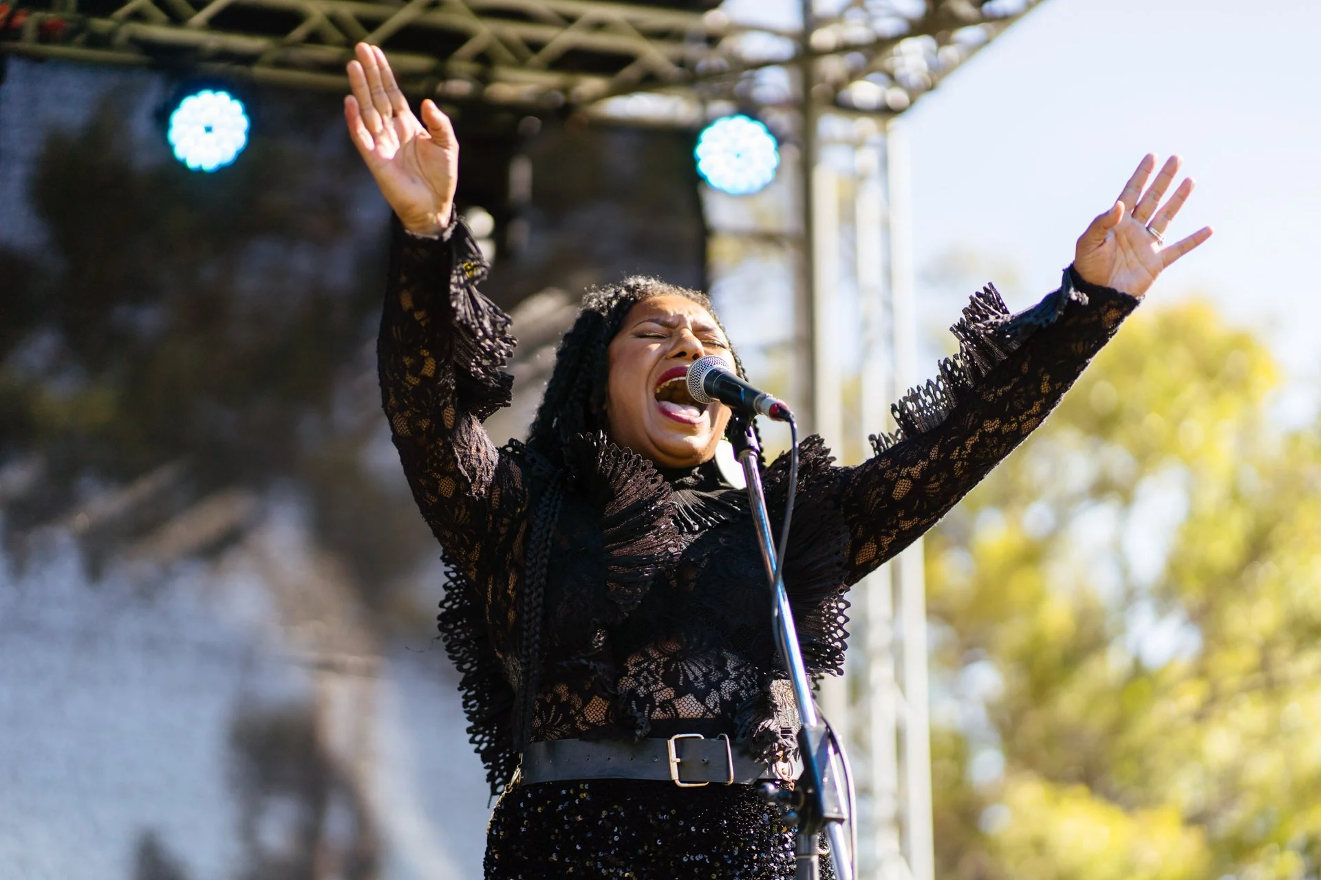 A woman singing passionately on stage with her arms raised, wearing a black lace outfit, with bright stage lights and trees in the background.