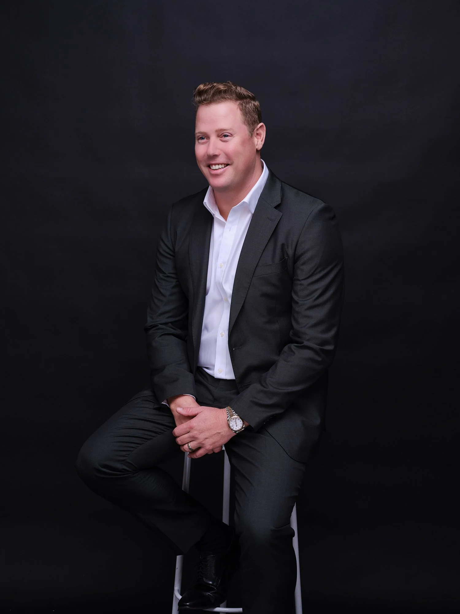 A man in a black suit white shirt sitting on a stool against a black background, smiling.