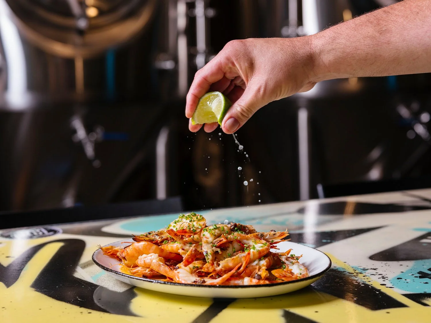 A hand squeezing lime over a plate of cooked shrimp and seafood garnished with herbs, on a colorful table.