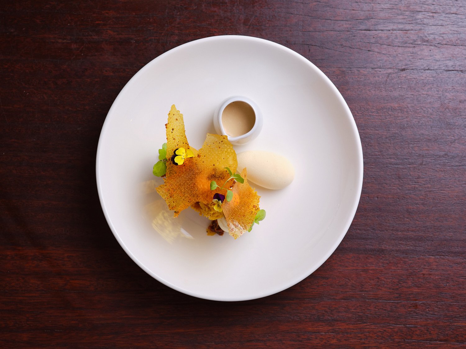 A white plate with a dessert featuring crispy lattice-like pastry, garnished with edible flowers and microgreens, served with a small container of sauce and a scoop of ice cream, all on a dark wooden table.