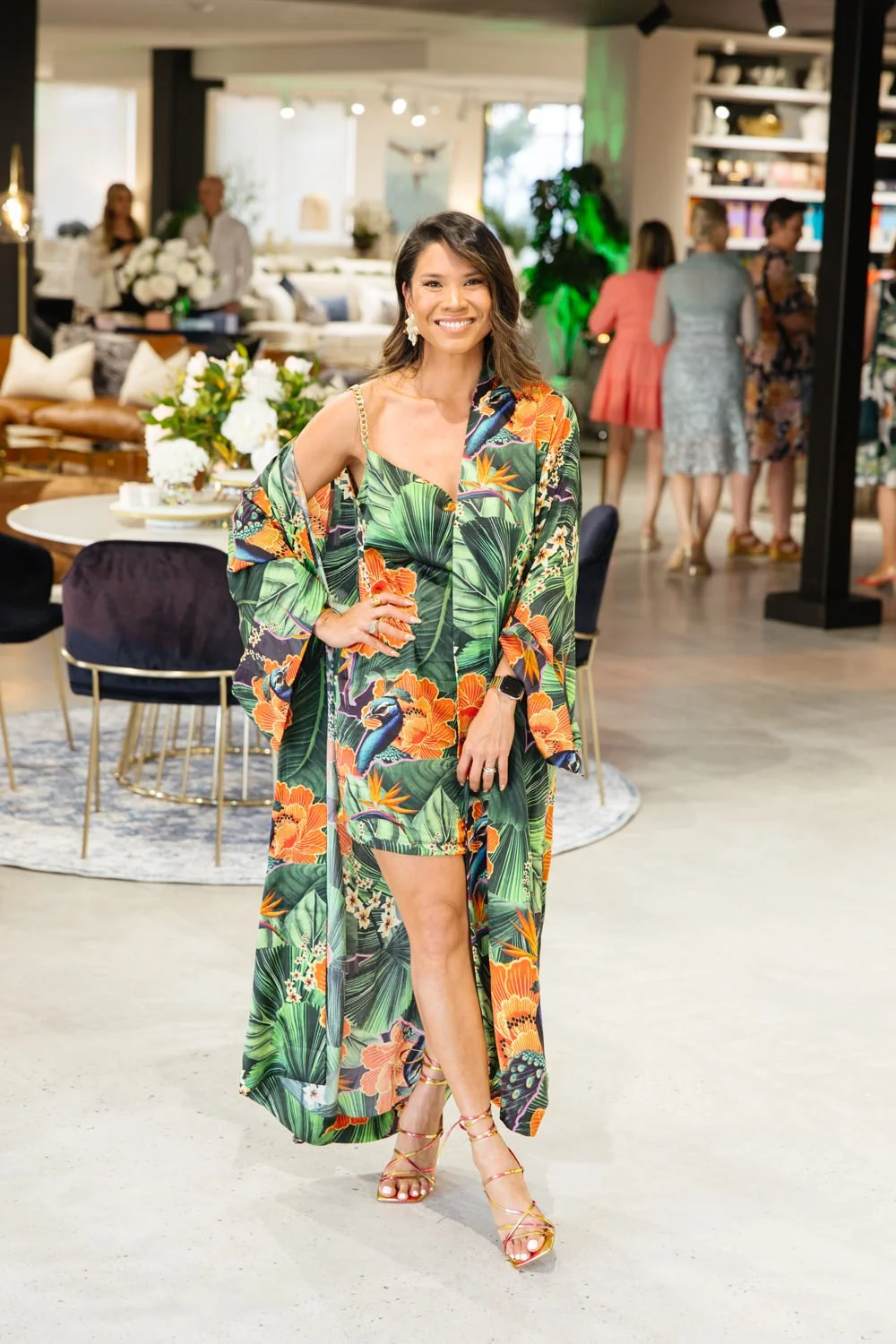 Woman in a colorful tropical dress and heels standing in a modern, well-lit indoor space with other women in the background.