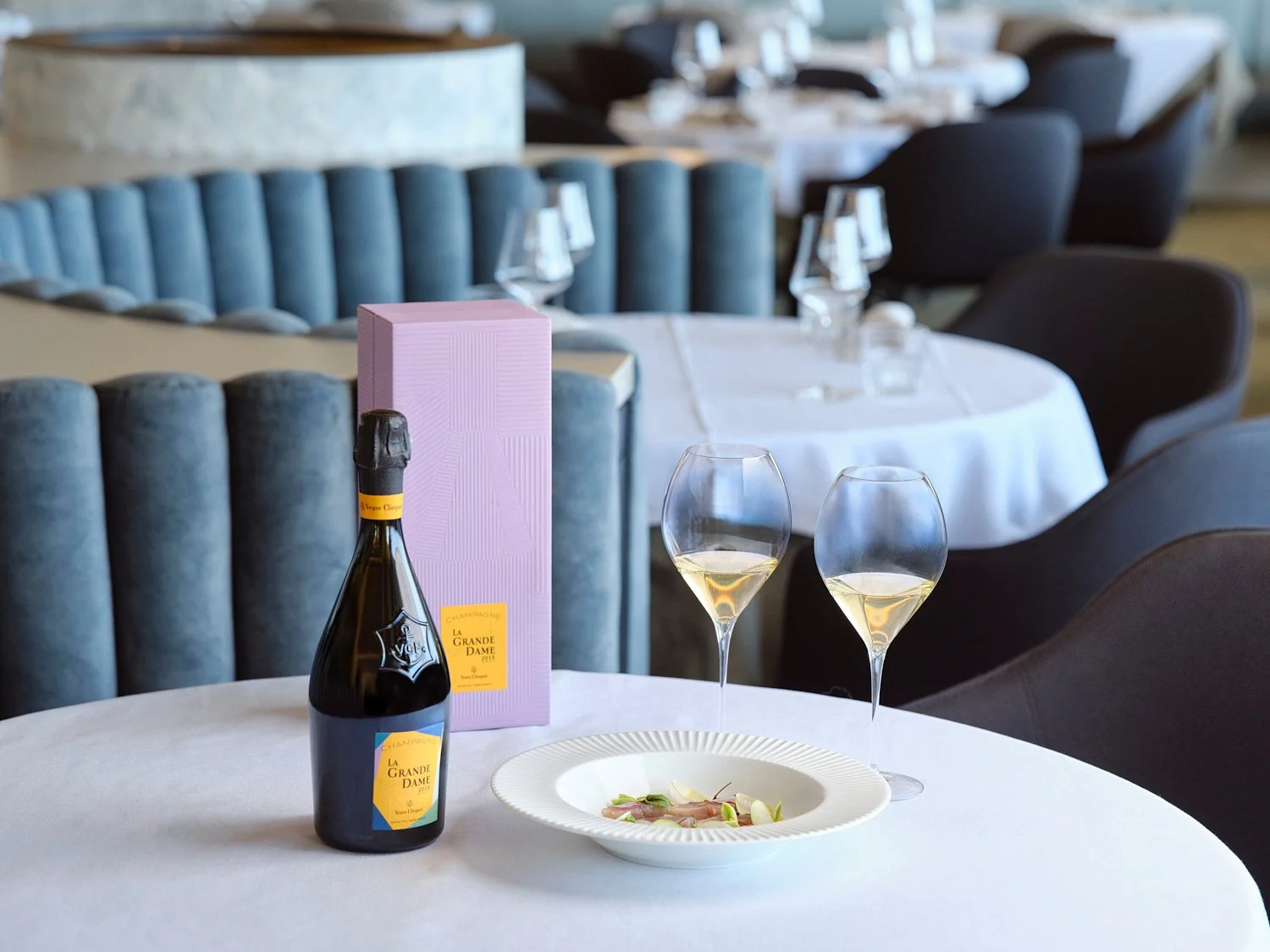A table set with a bottle of La Grande Dame champagne, two glasses filled with champagne, a white plate with food, and a pink gift box in a restaurant with white tablecloths and elegant chairs.