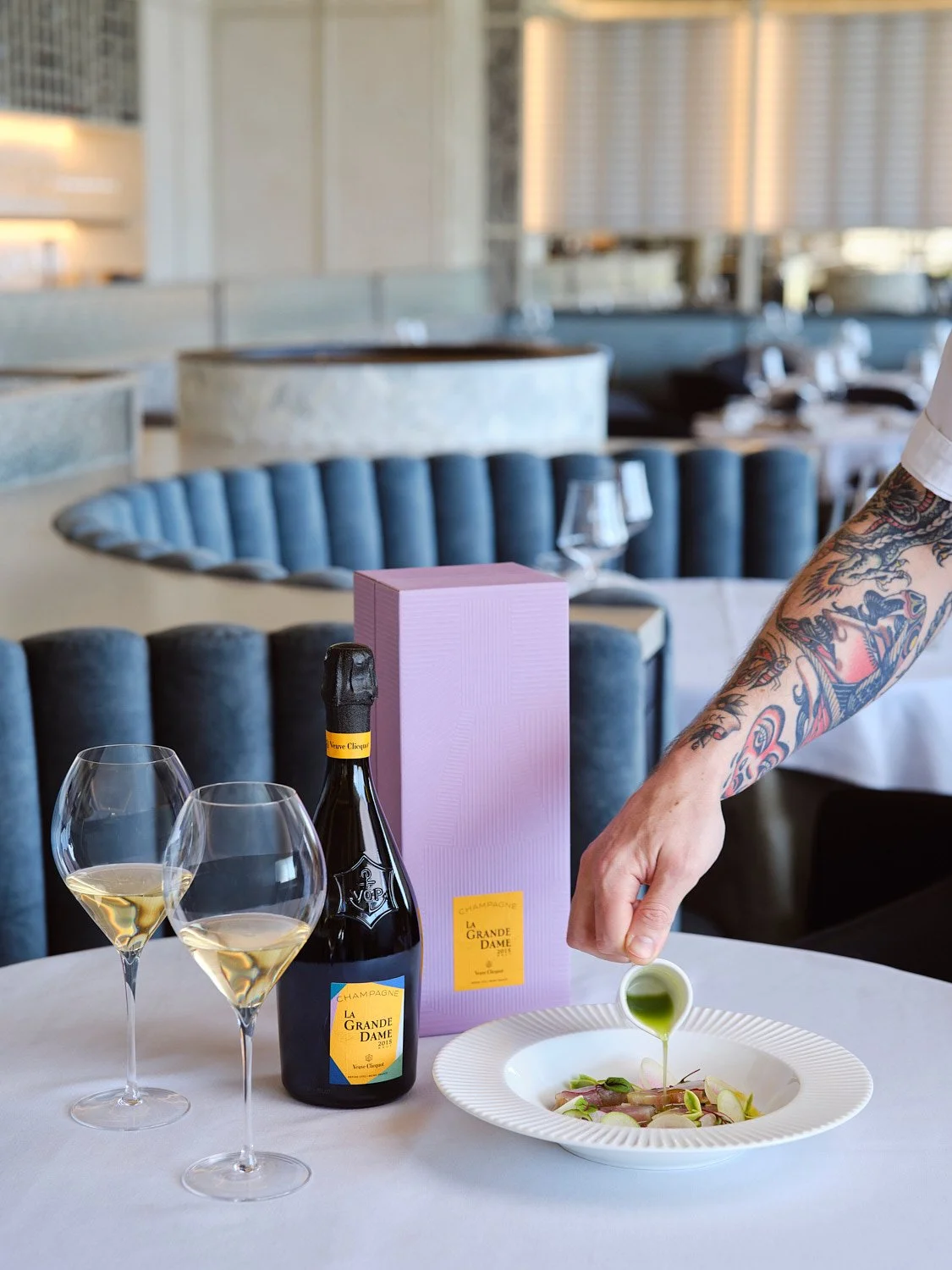 A person with a tattooed arm pouring green sauce onto a dish with sliced vegetables at a restaurant table set with a bottle of La Grande Dame champagne, two filled champagne glasses, and a purple gift box.