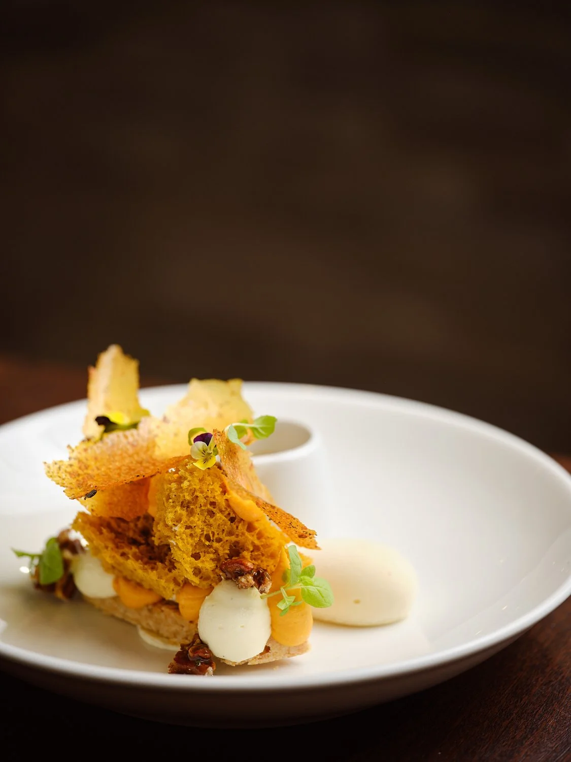 A plated fine dining dish with crispy bread, microgreens, dollops of white and orange sauces, and garnishes on a white plate.