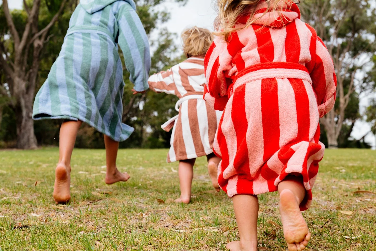 Three children wearing striped bathrobes running barefoot on grass outdoors, with trees in the background.