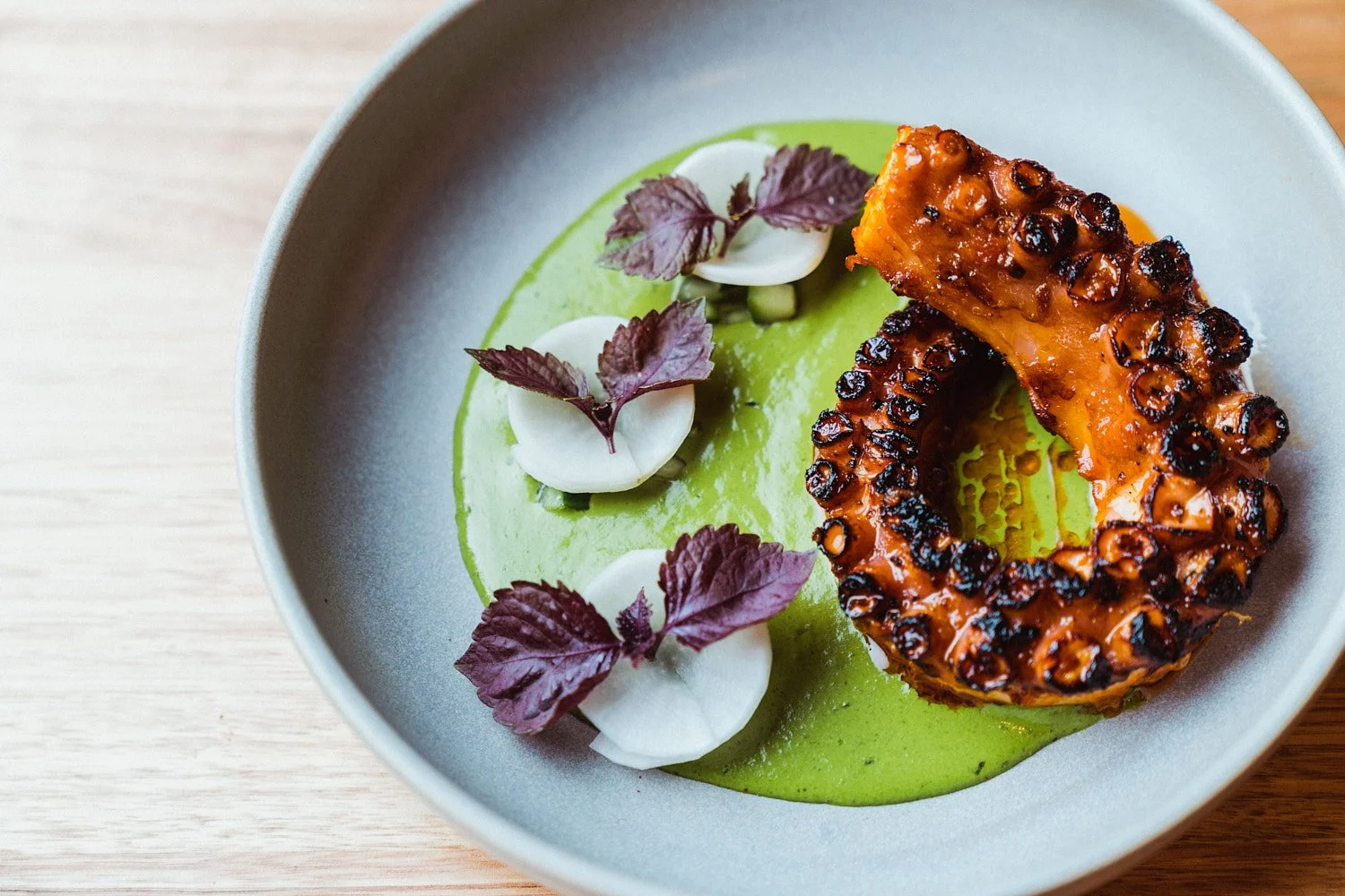 Grilled octopus tentacle served on a gray plate with green sauce, garnished with small purple leaves and dollops of white sauce.