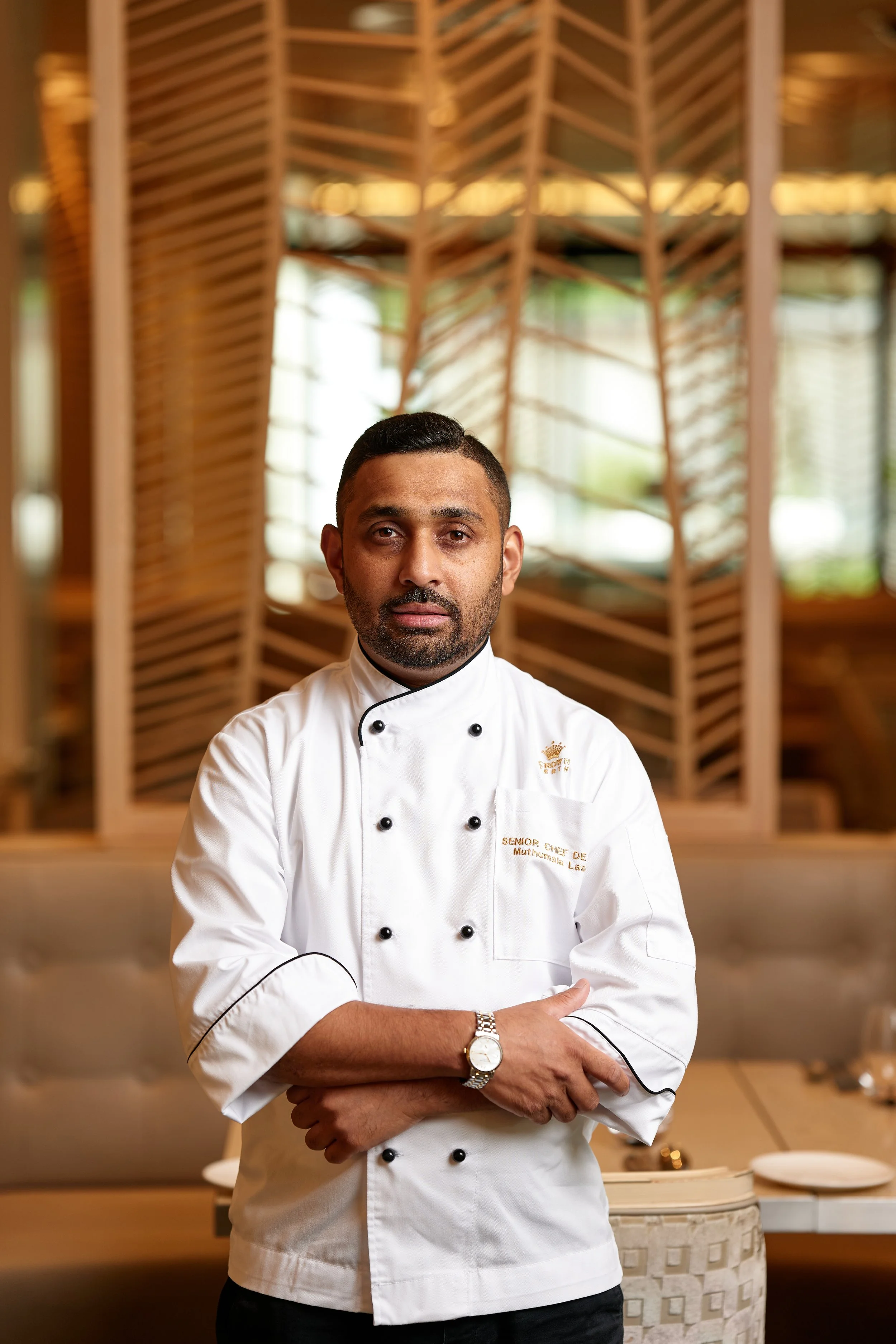 A male chef in a white chef's coat with black buttons standing with crossed arms in a restaurant dining area.