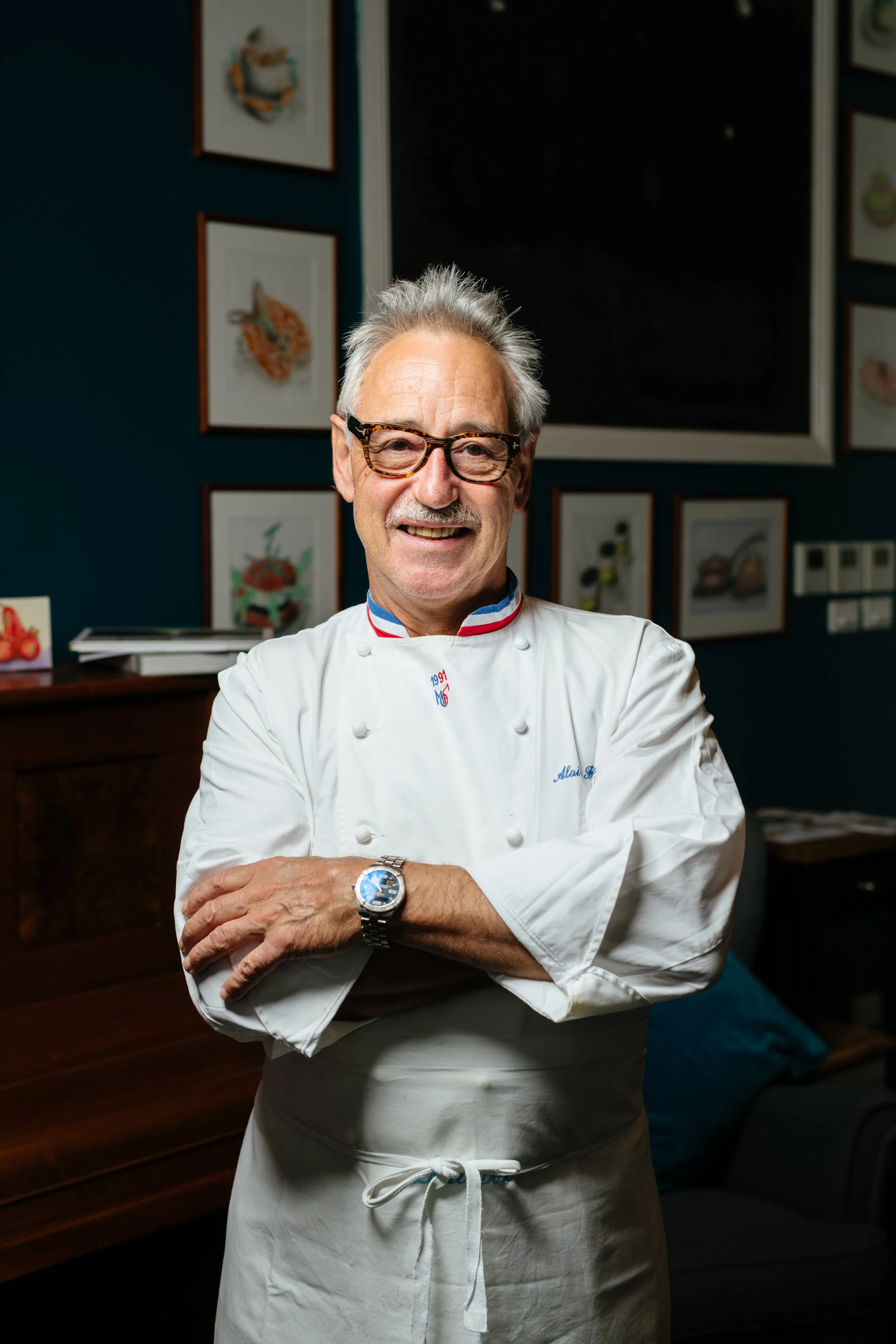 A smiling chef with gray hair and glasses, wearing a white chef’s coat and apron, standing with arms crossed in a restaurant or kitchen setting, with framed food artwork on the wall behind him.