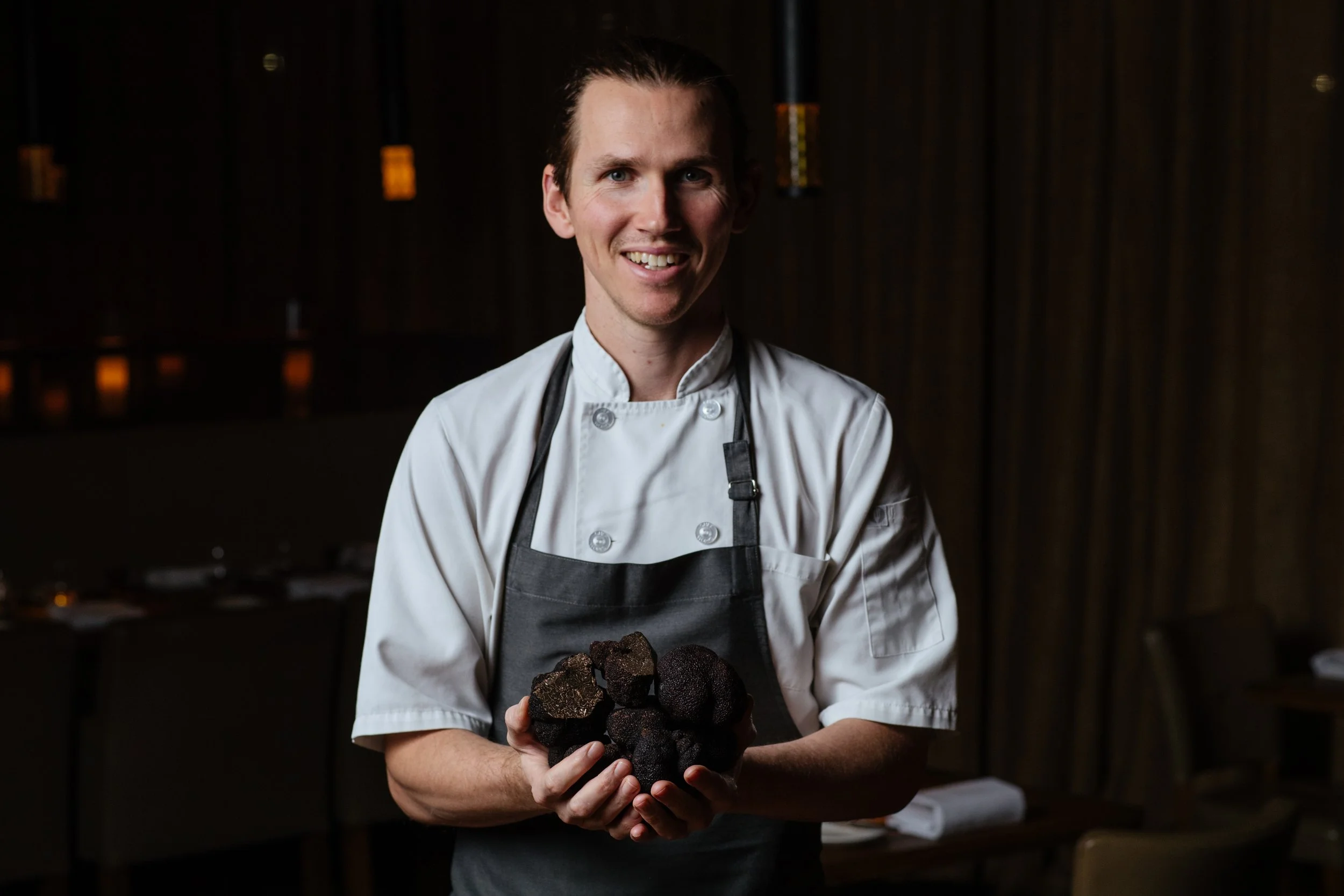 Chef smiling and holding a cluster of black truffles in a dimly lit restaurant setting.