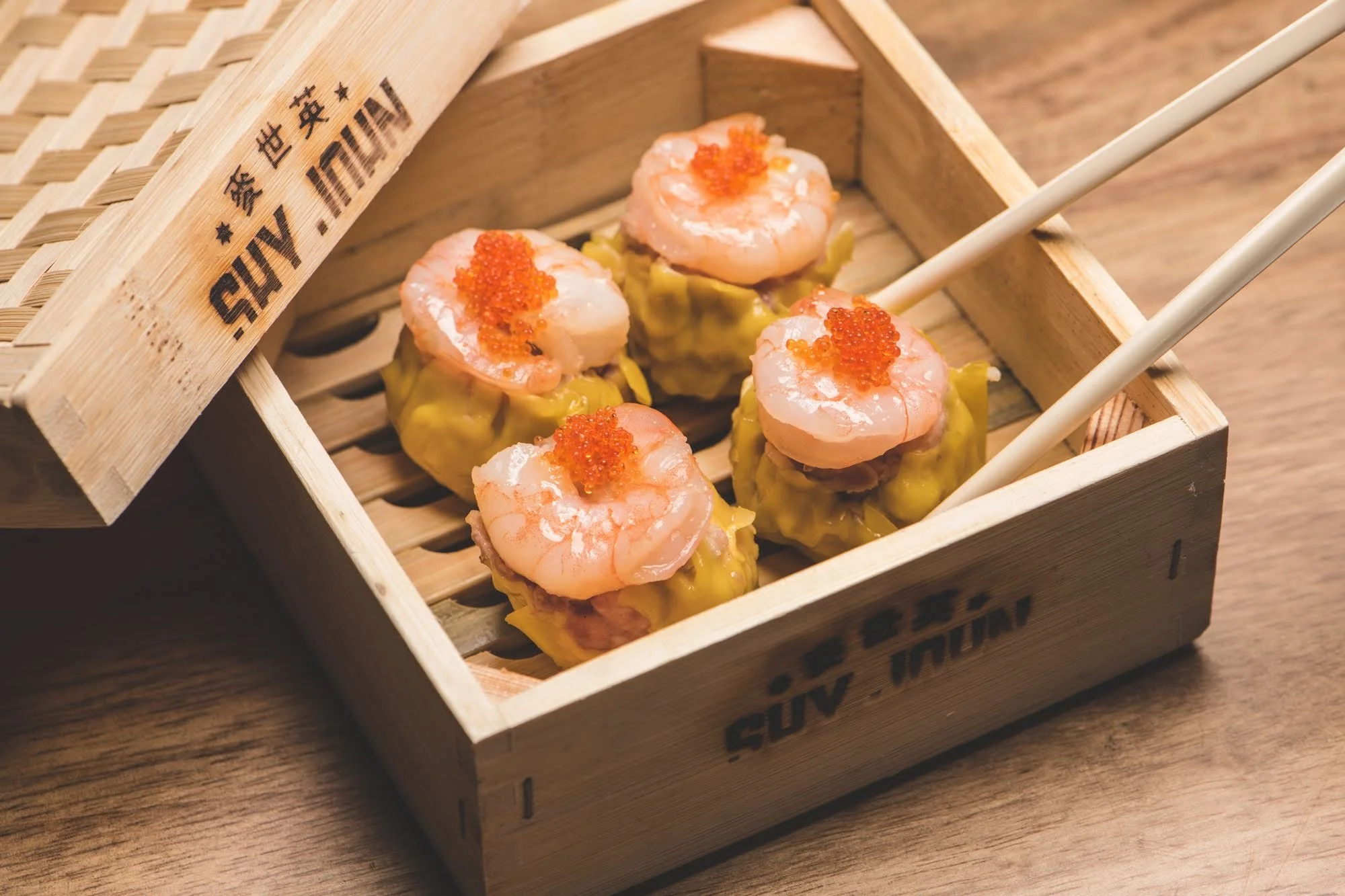Four pieces of shrimp and fish shumai with orange roe on top, served in a wooden box with chopsticks.