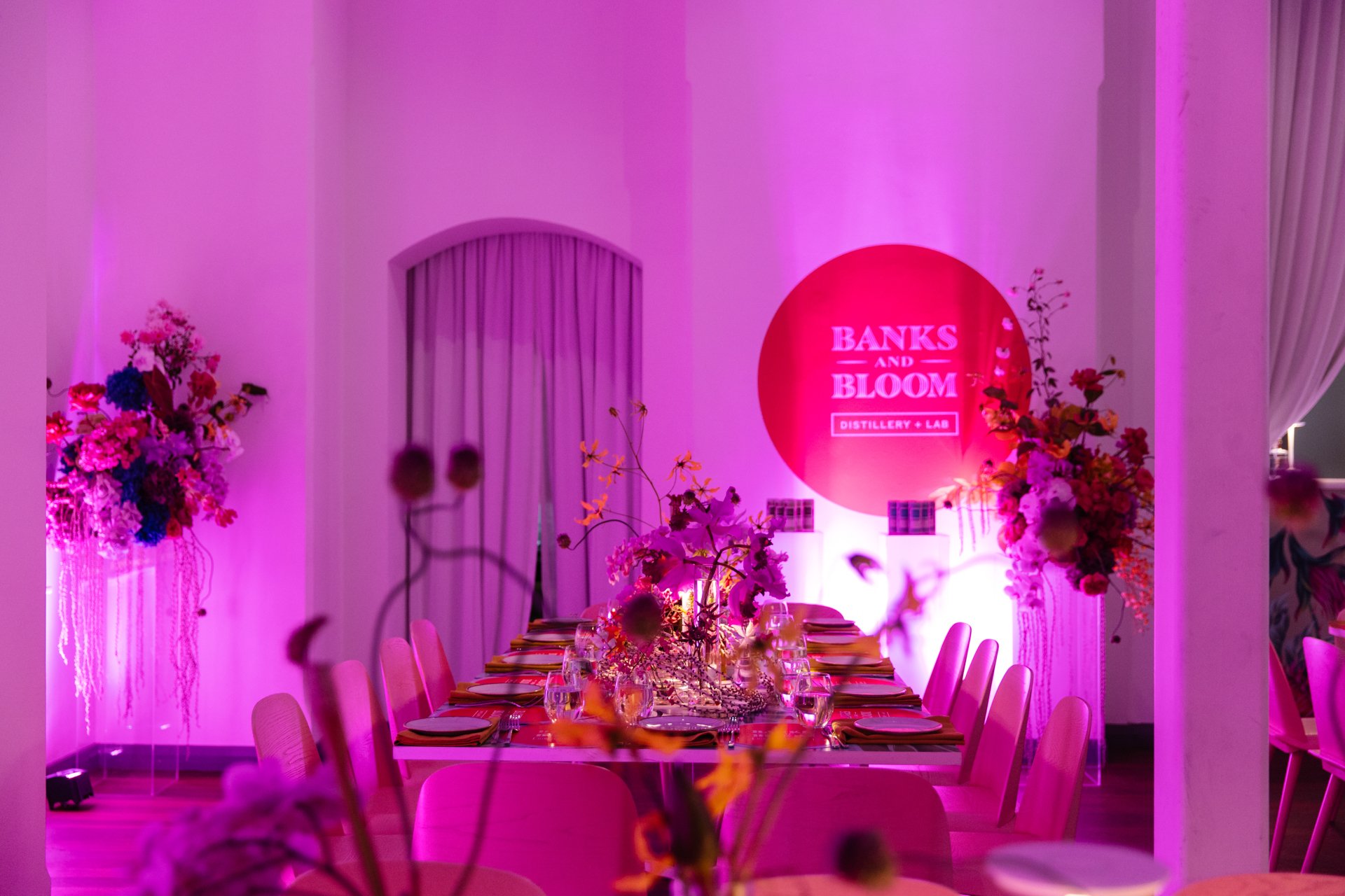 A dinner event setup with a long table decorated with flowers under pink and purple lighting, with a sign that reads 'Banks and Bloom Distillery + Lab' on the wall.