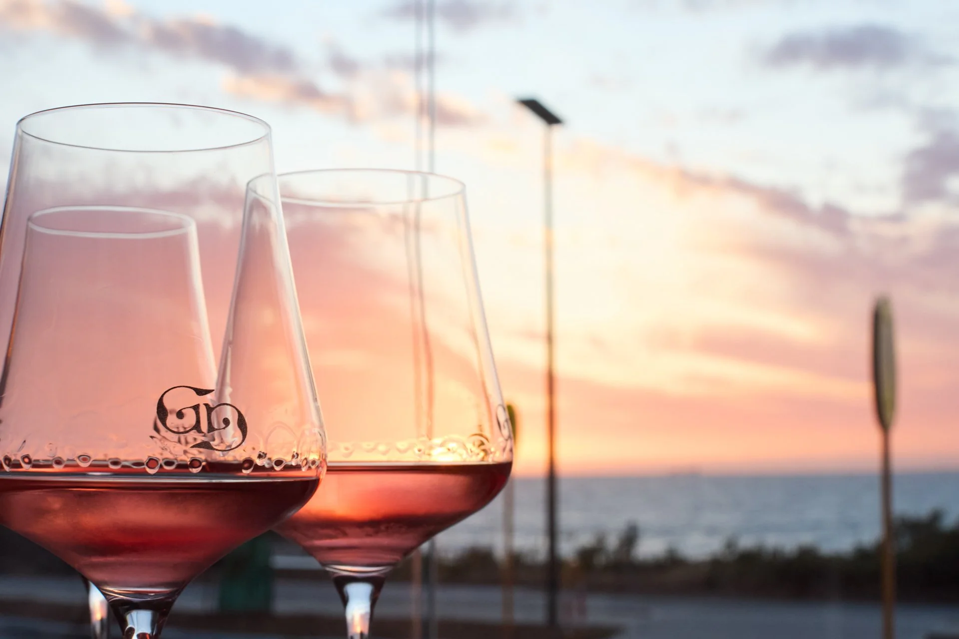 Three glasses of pink wine in front of a sunset over the ocean with palm trees and a pier in the background.