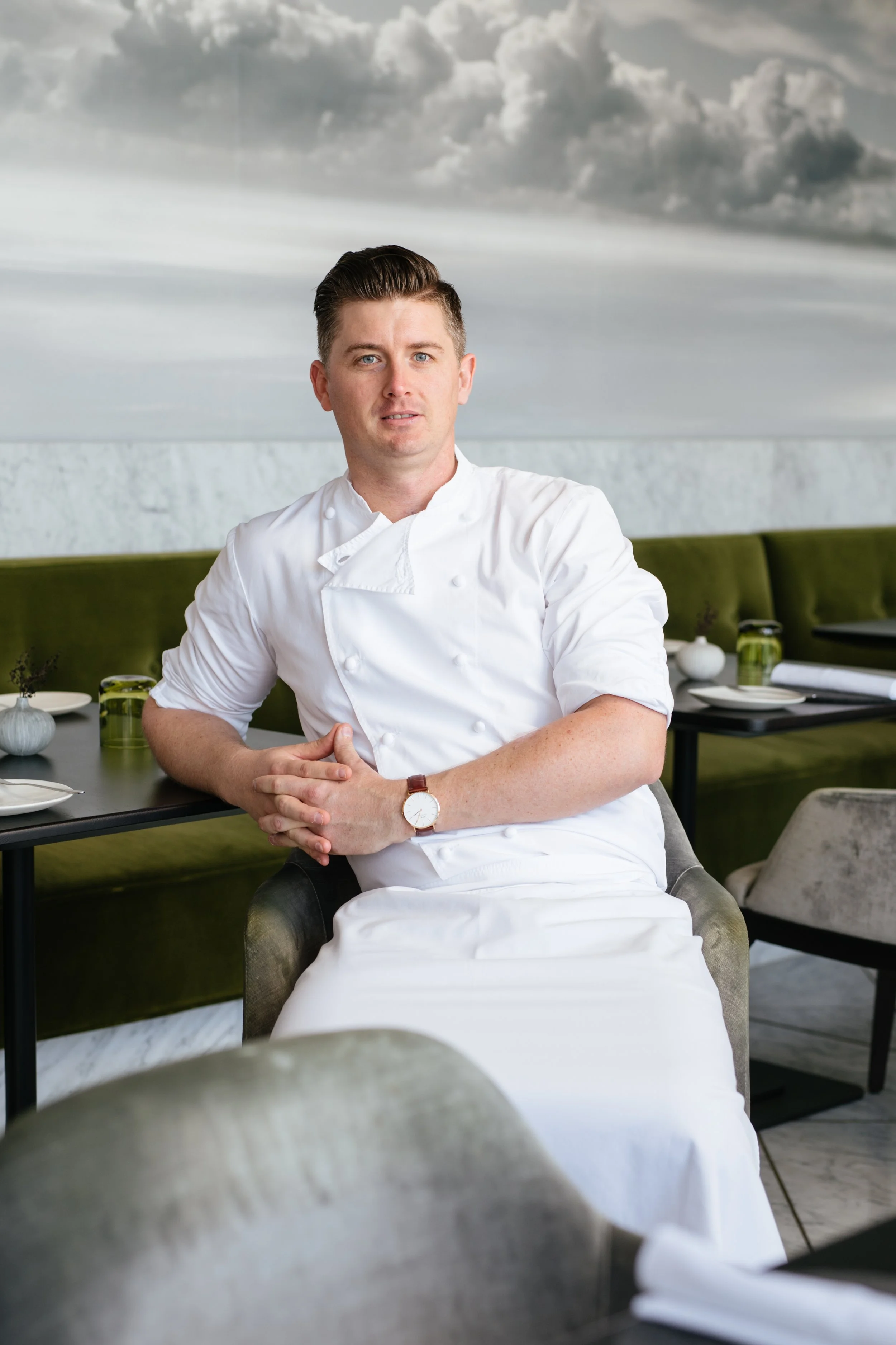 A male chef in a white chef's uniform sitting at a table in a restaurant, with a modern interior and a large cloud mural on the wall behind him.