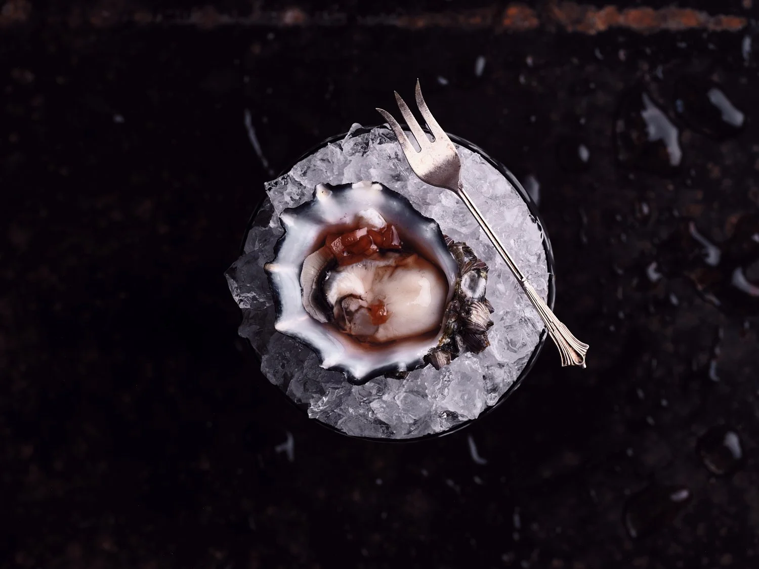 A fresh oyster on a bed of ice in a glass, with a small fork resting on top, on a dark surface.