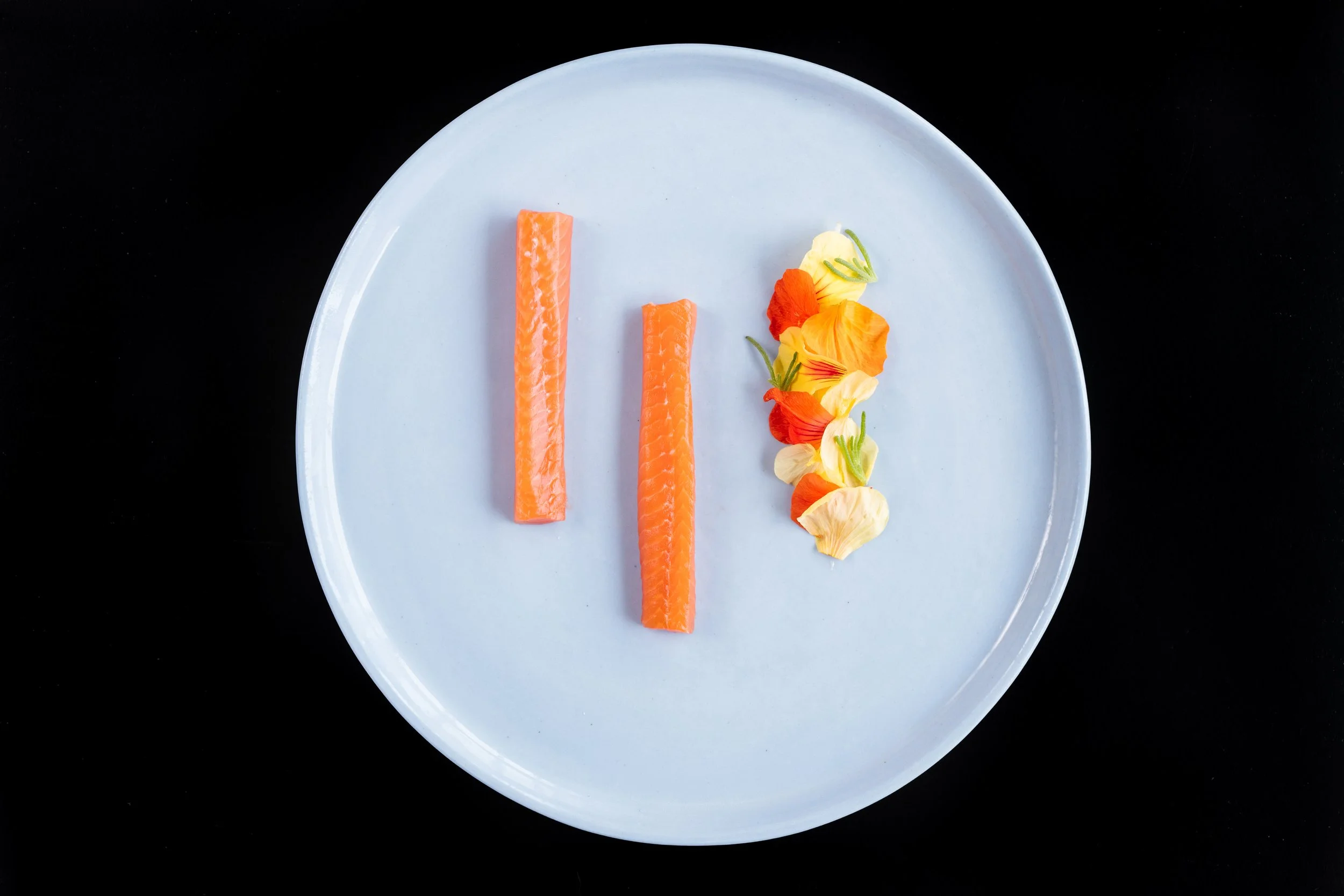 Two pieces of raw salmon sashimi and edible flower petals on a white plate against a black background.