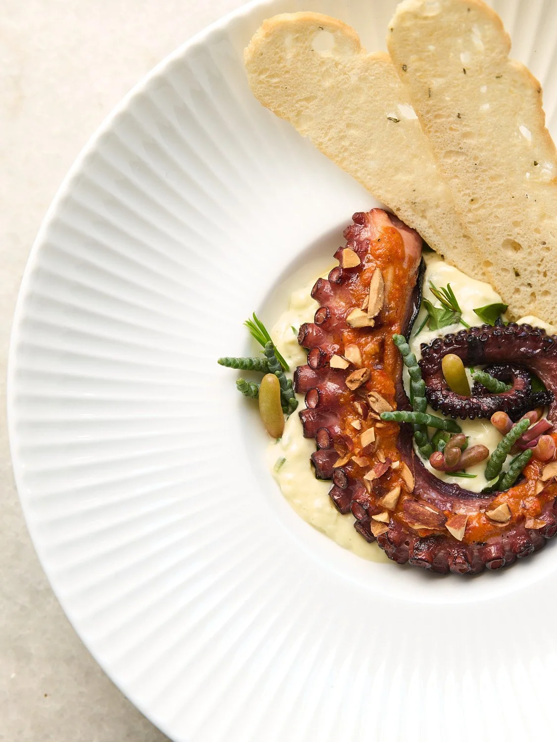 Grilled octopus tentacle served on a bed of white sauce with sliced almonds, garnished with herbs and paired with two slices of toasted bread on a white plate.