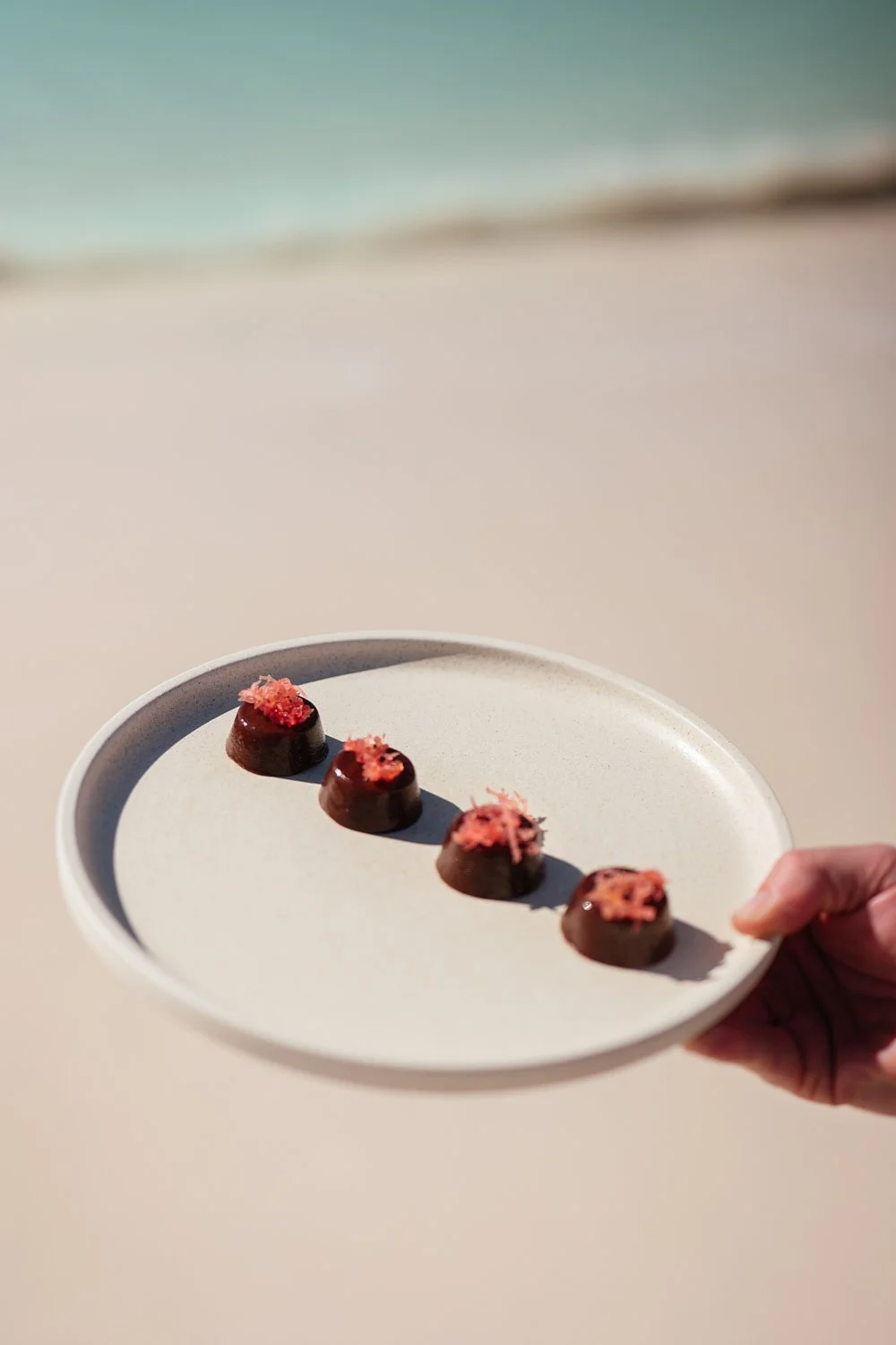 A hand holding a white plate with four small chocolate desserts topped with pink decorations, against a blurred beach background.