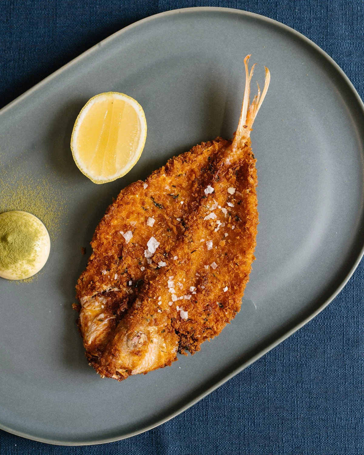 Crispy fried fish fillet on a gray plate with a lemon wedge and a dollop of green sauce, on a dark blue tablecloth.