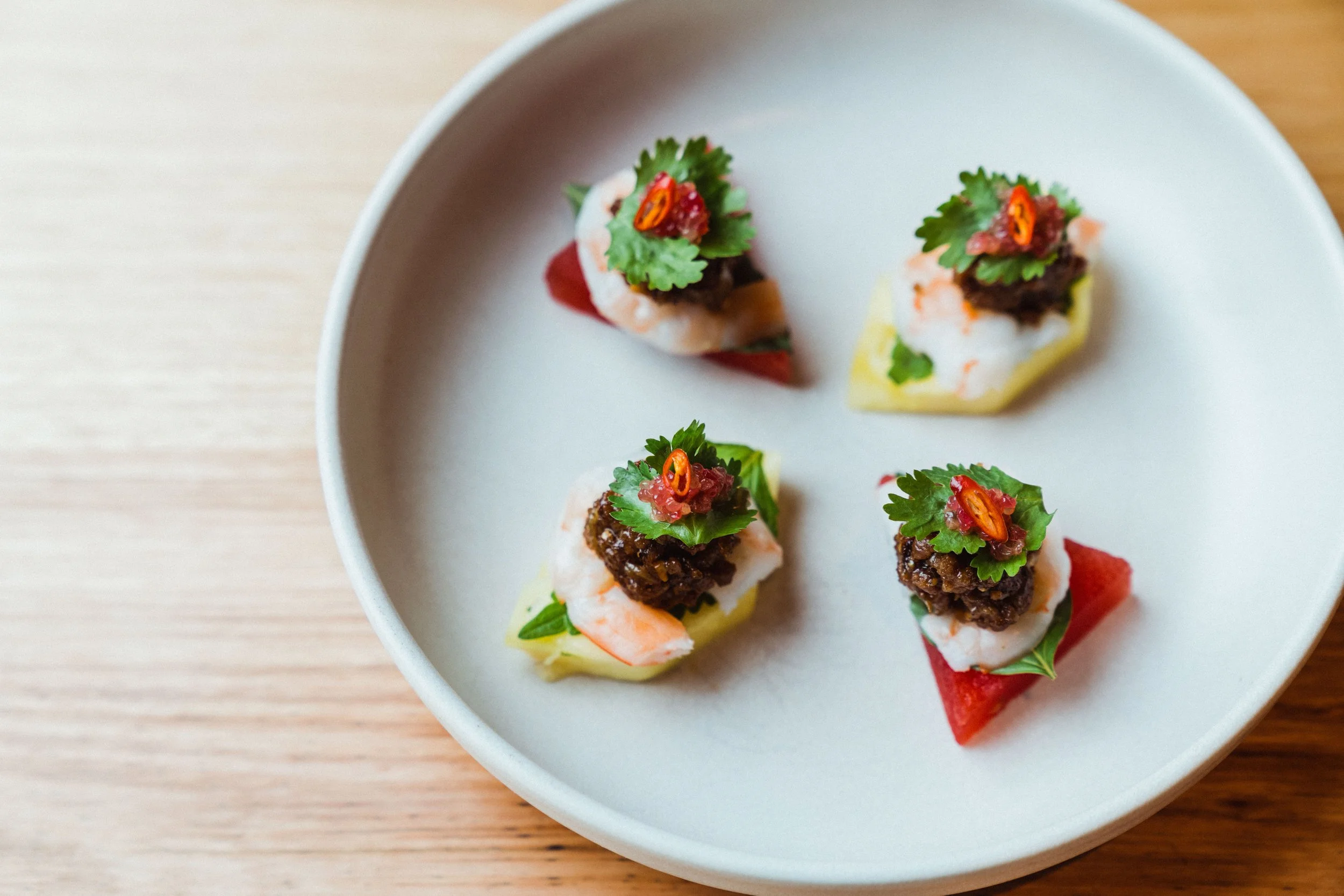 Assorted bite-sized appetizers topped with herbs and chili, on a white plate.