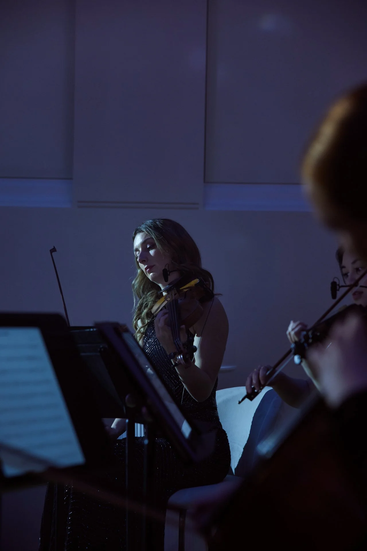 A young woman playing the violin during a performance, illuminated by soft stage lighting, with other musicians partially visible in the background.