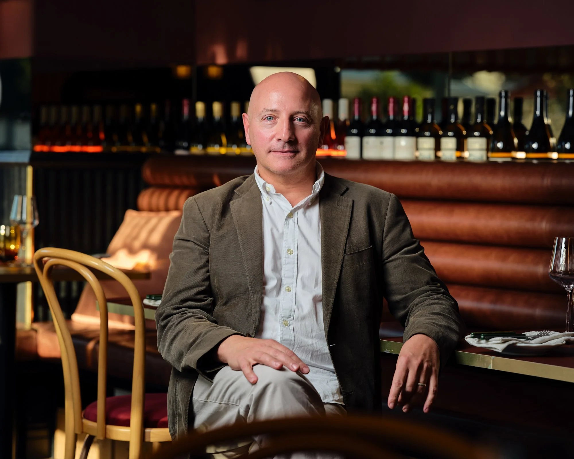 A man with a bald head and light skin sitting at a table in a dimly lit restaurant with wine bottles on a shelf behind him, wearing a white shirt and a gray blazer.