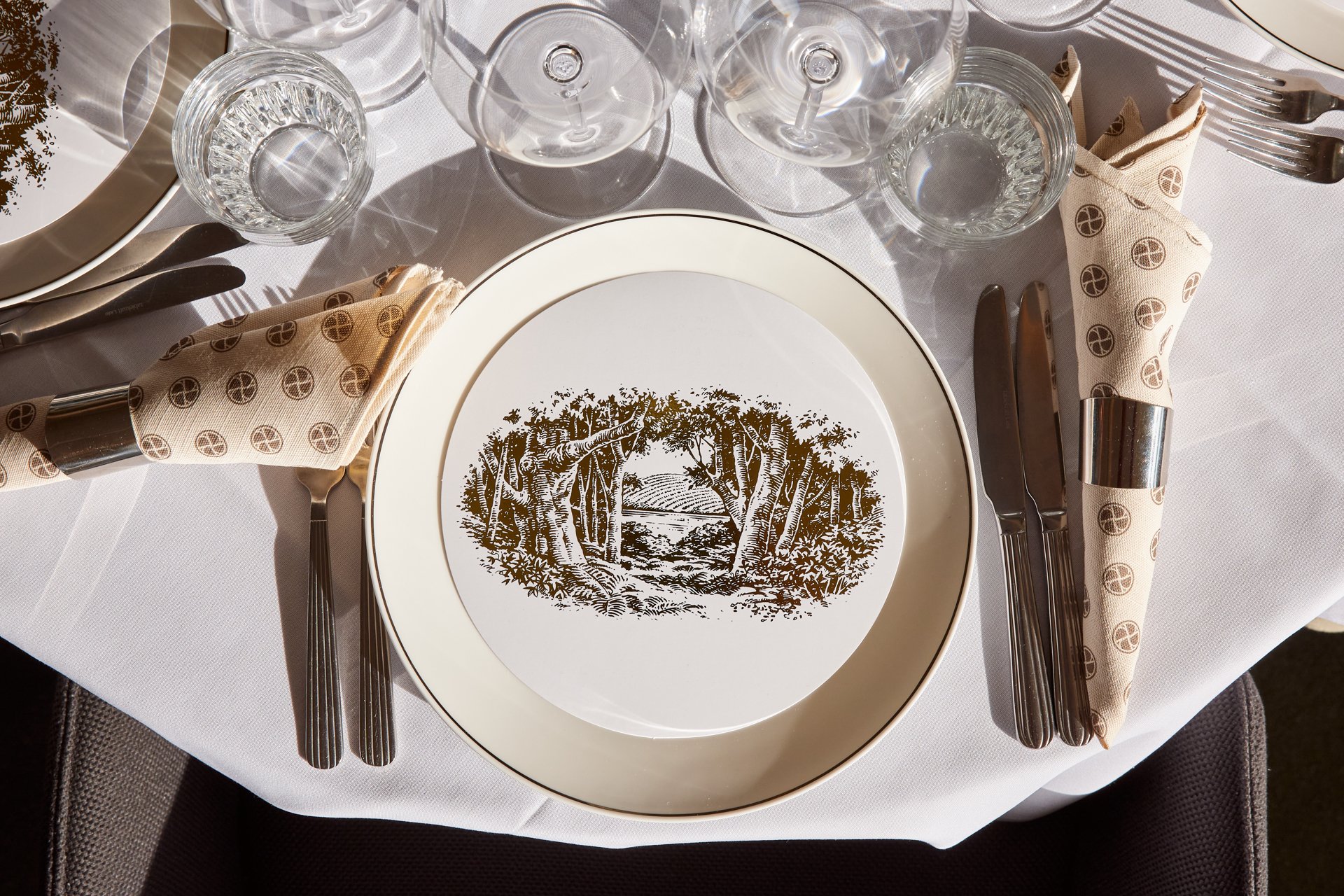 Elegant dining table setting with a decorative plate featuring a black and white illustration of a forest scene, cream napkins with a circular pattern, glassware, and silverware on a white tablecloth.