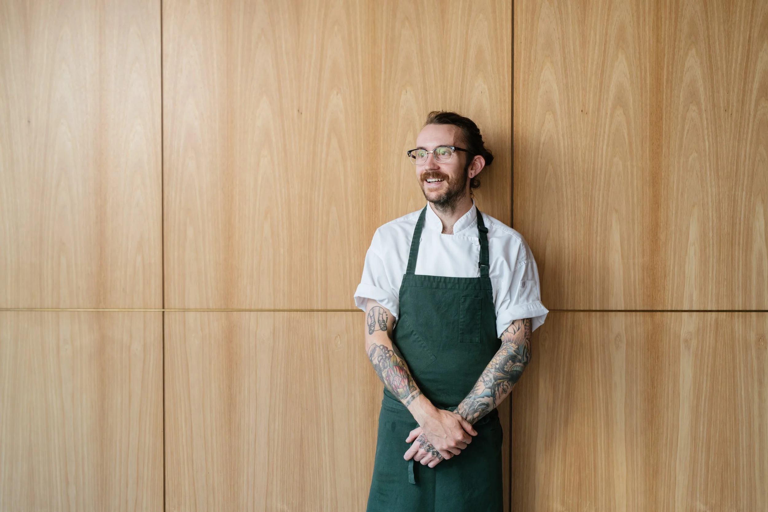 A man wearing glasses, a white chef's coat, and a green apron, standing against a wooden wall with his arms crossed and smiling.