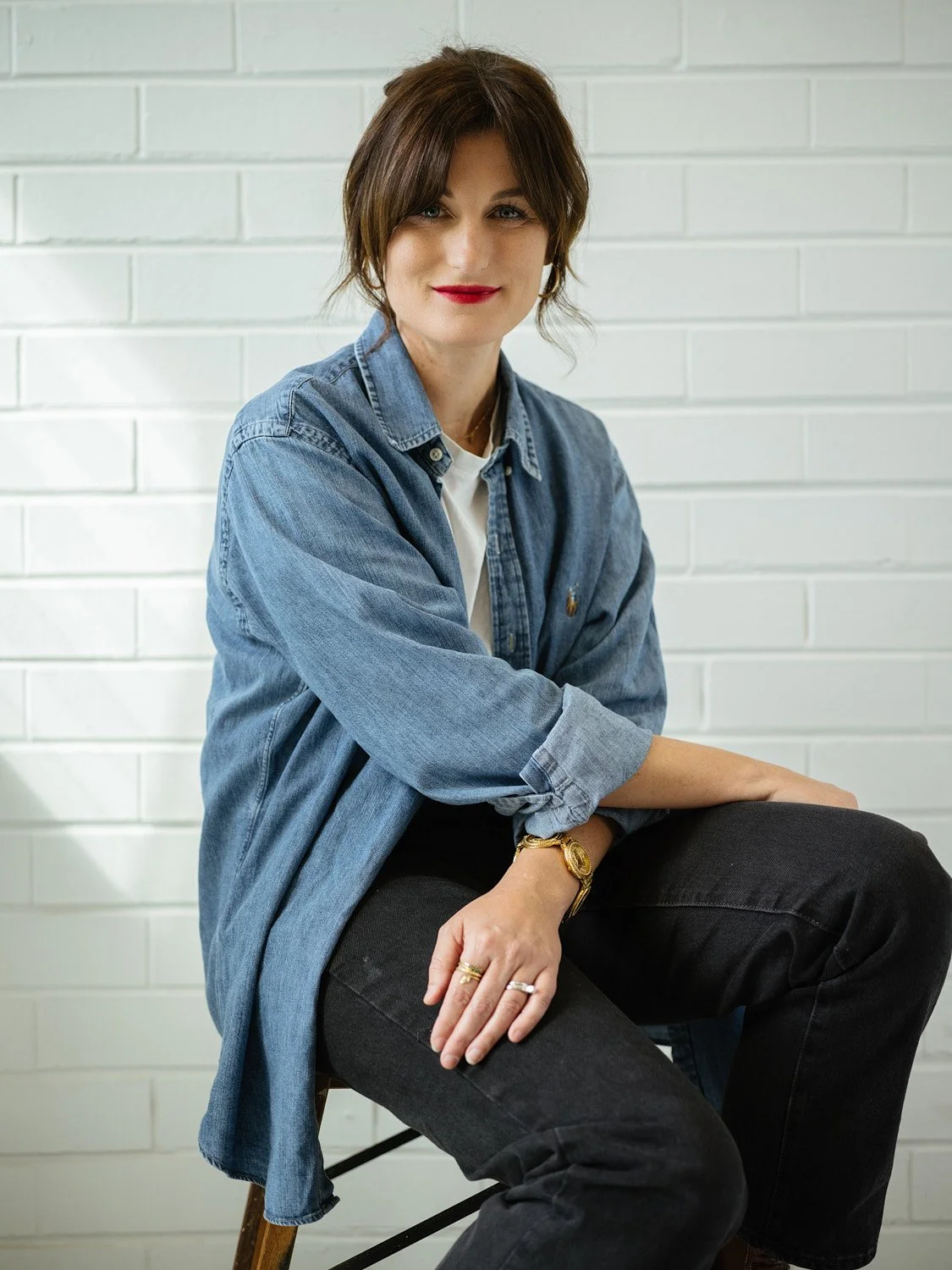 A woman with short brown hair and red lipstick wearing a denim jacket, sitting against a white brick wall.