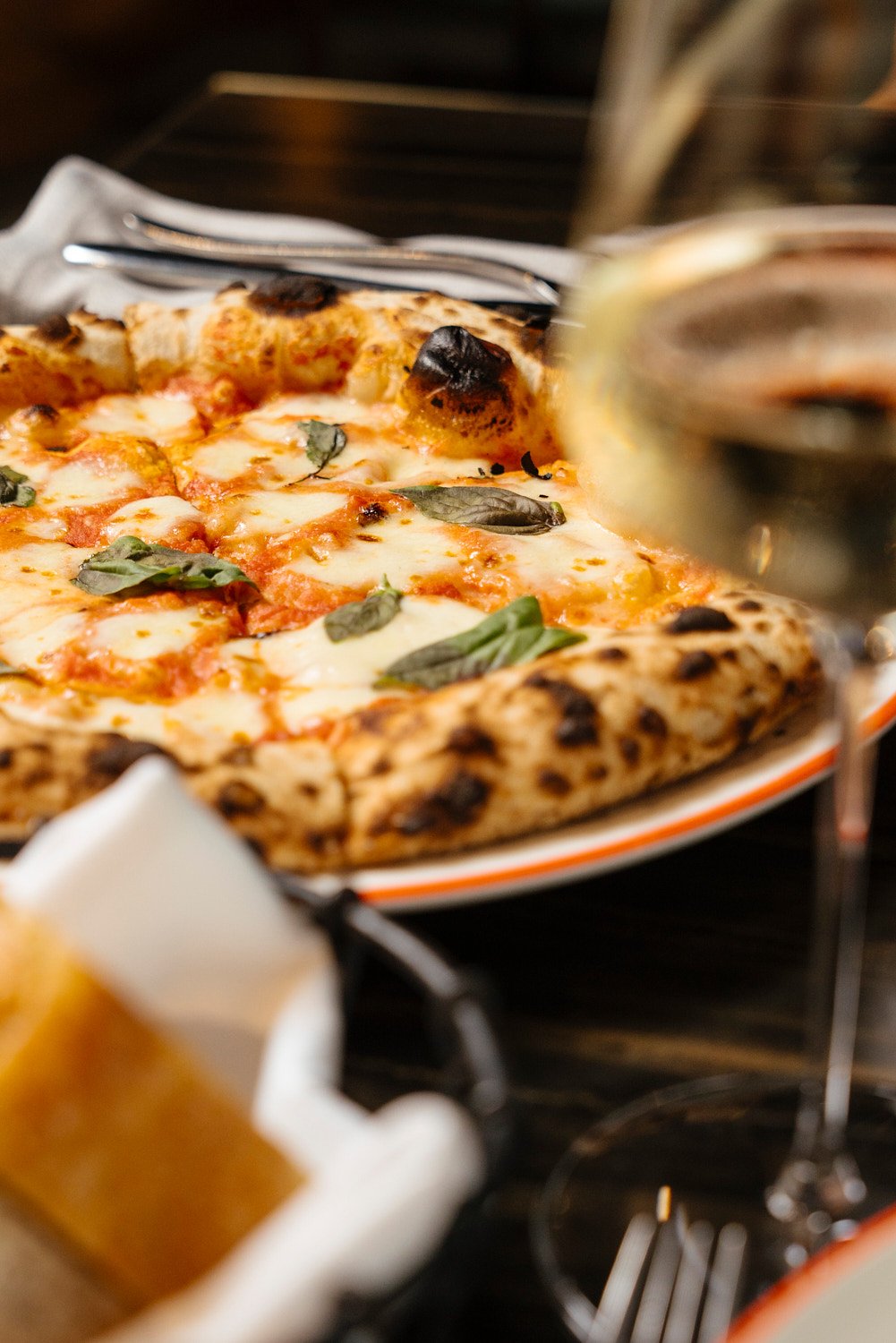 Close-up of a freshly baked Margherita pizza with melted cheese, basil leaves, and a slightly charred crust on a plate, with a glass of white wine nearby.