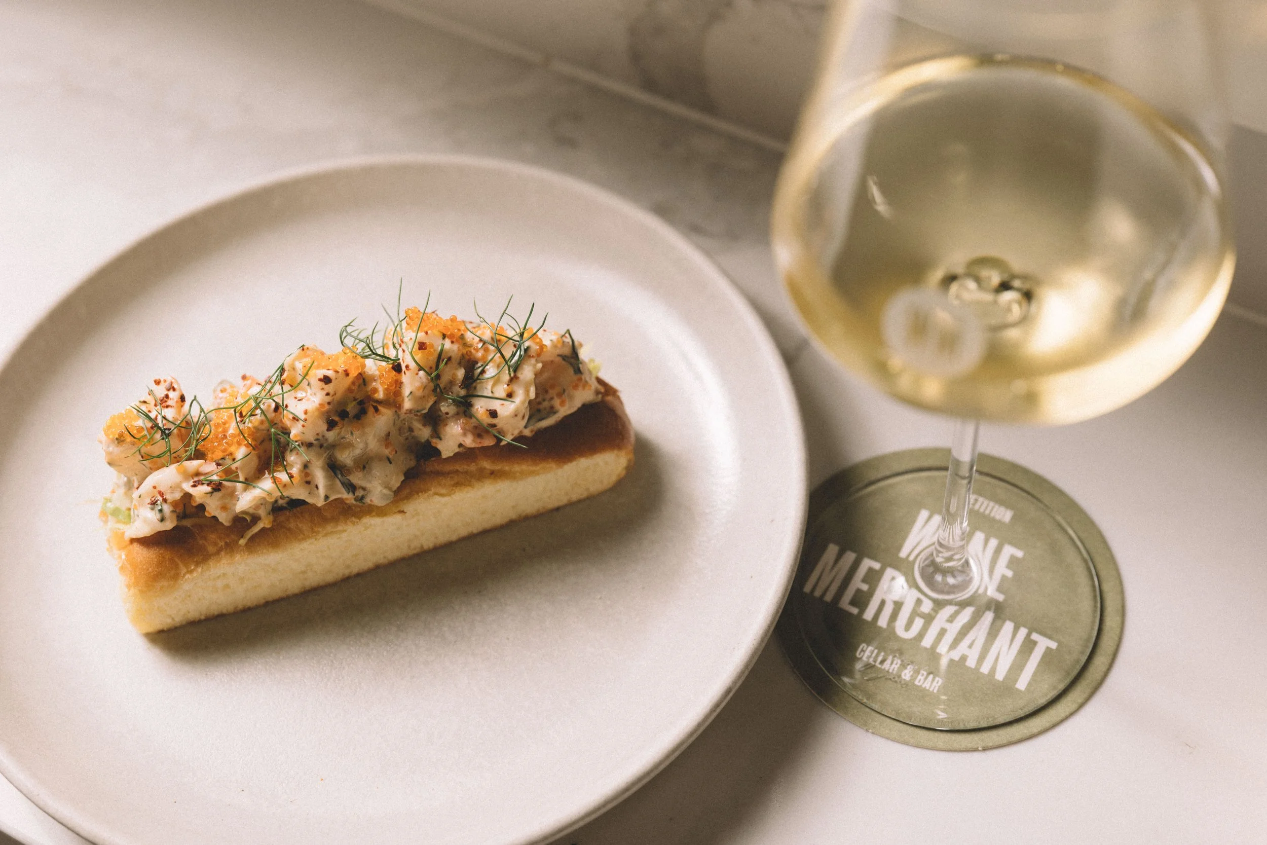 A slice of savory dish with diced seafood, garnished with herbs, served on a white plate, with a glass of white wine on a bar coaster labeled 'Wine & Mercant'.