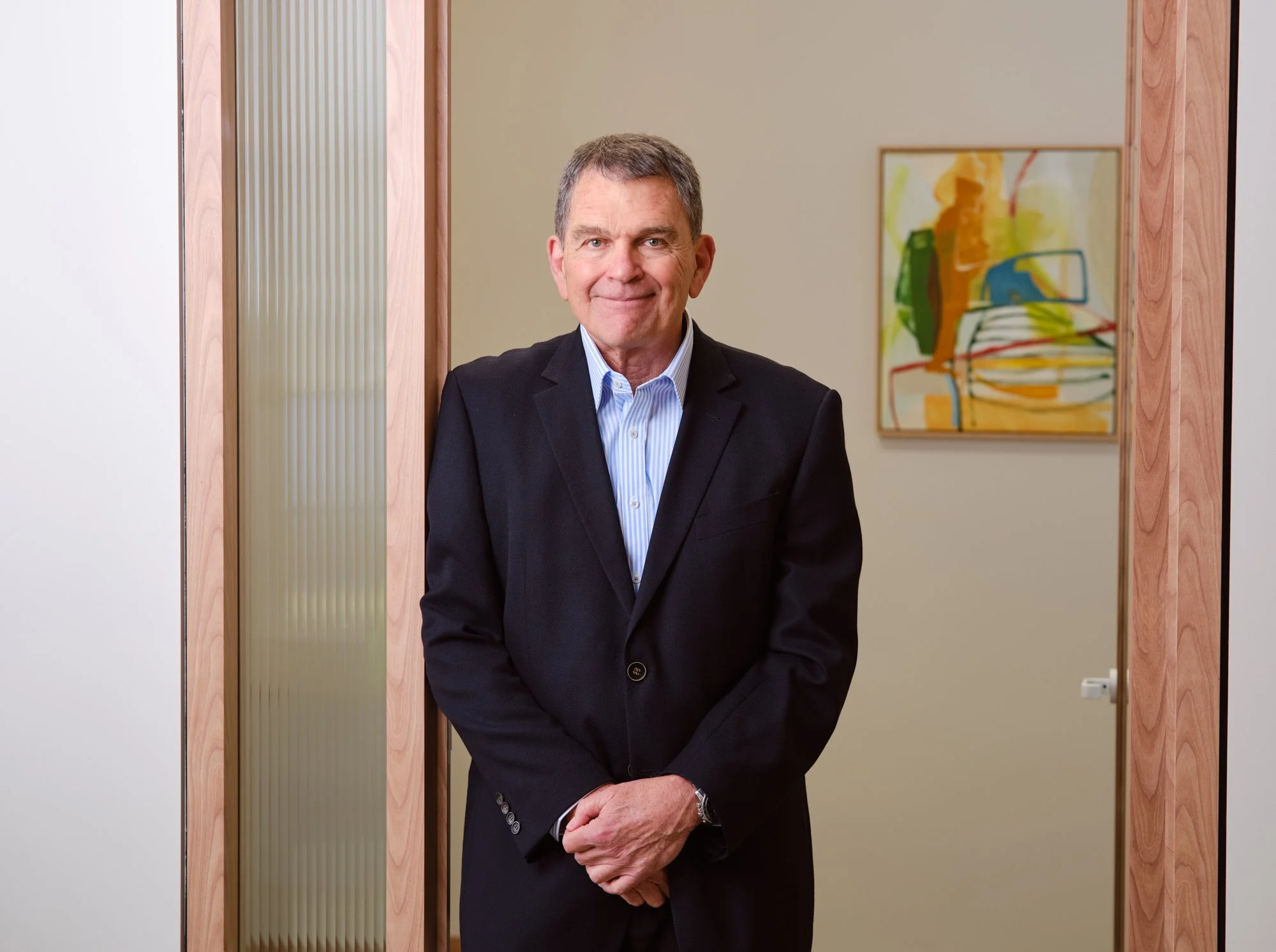 A middle-aged man in a black suit and light blue shirt standing inside an office doorway.