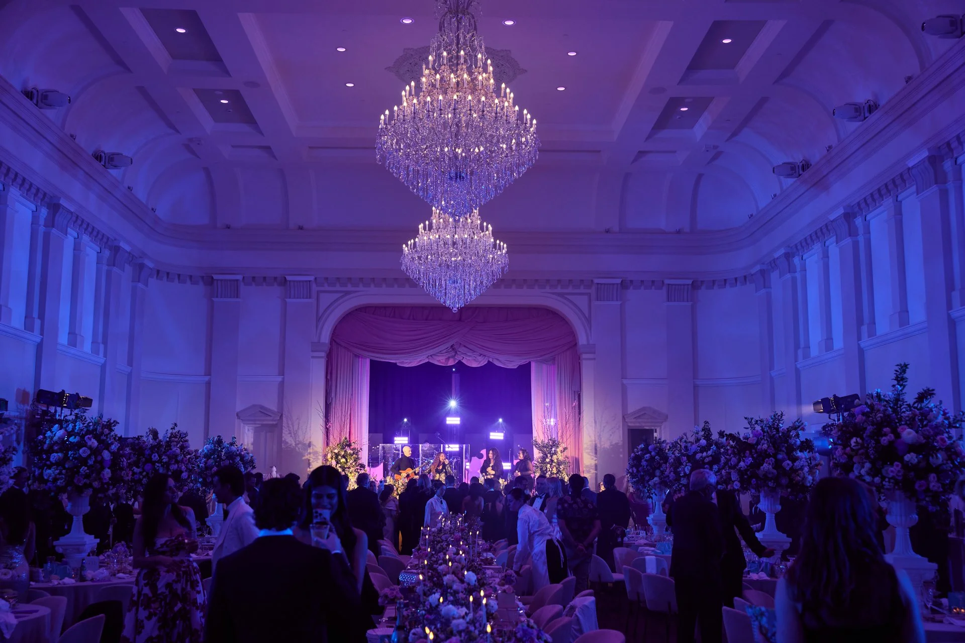 Elegant event hall with large chandeliers, purple lighting, floral arrangements, and a stage with live band performers, surrounded by guests