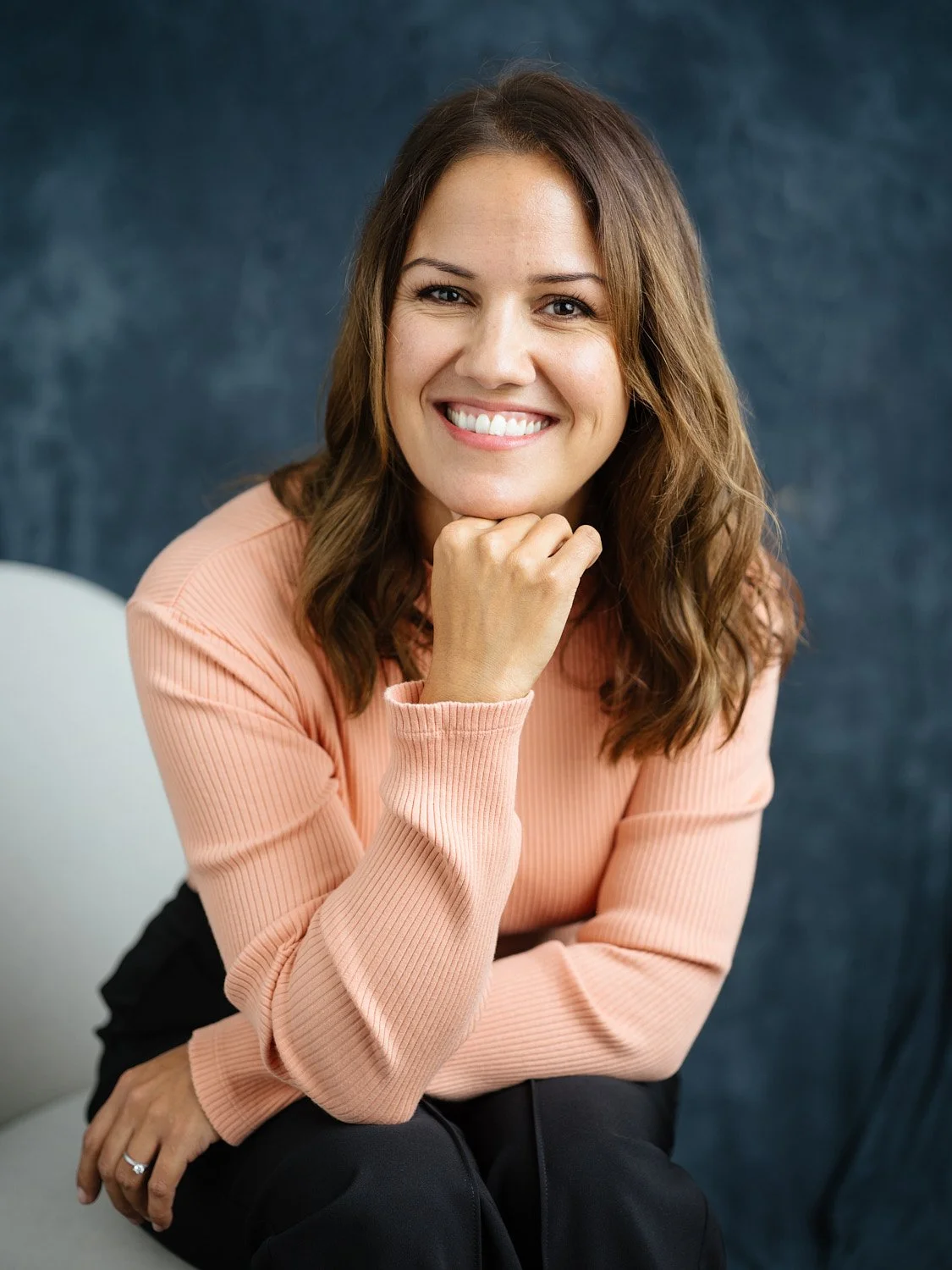 A smiling woman with shoulder-length brown hair, wearing a peach-colored sweater, sits with her chin resting on her hand against a dark blue textured background.