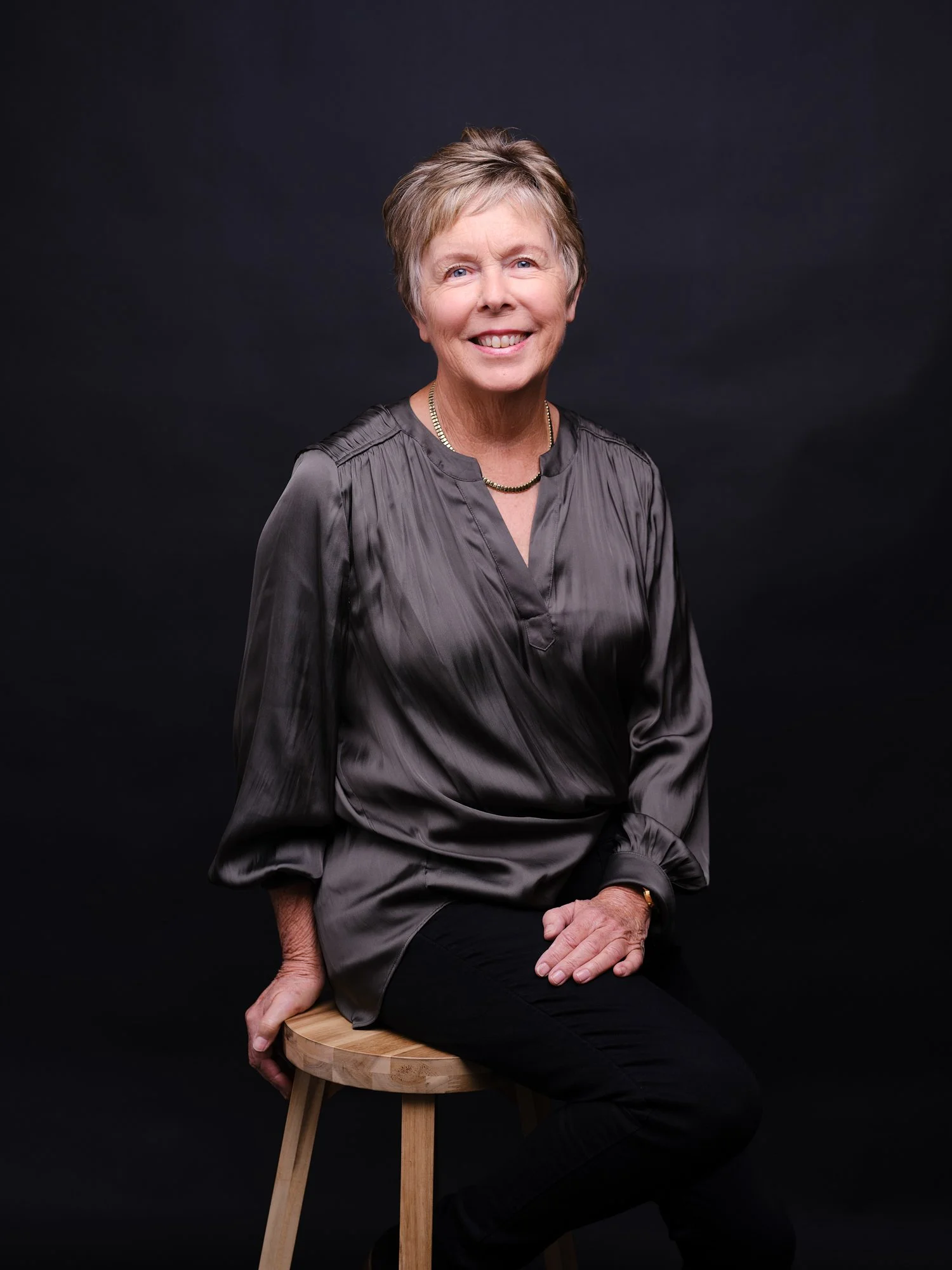 A smiling elderly woman with short light brown hair, wearing a shiny gray blouse and black pants, sitting on a wooden stool against a plain black background.