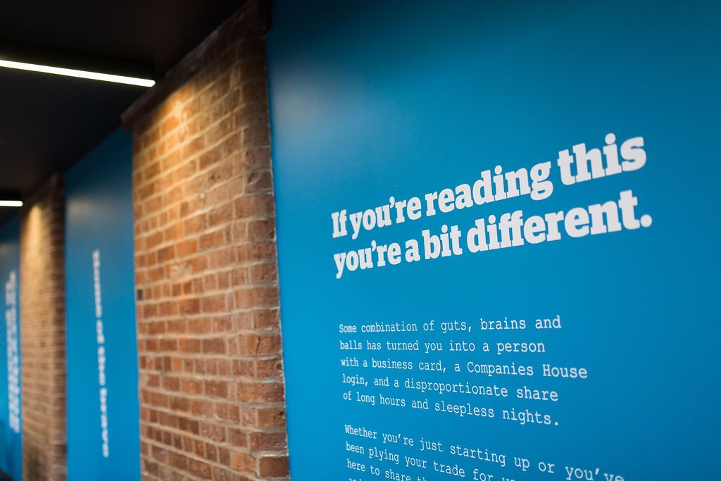Bold branded wall graphics within a heritage mill office refurbishment, combining exposed brickwork and contemporary workplace design to create an inspiring commercial interior by Stiwdio Interior Design, North Wales.