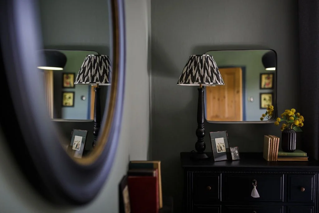 A black table with framed photos, a black lamp with a patterned shade, and a vase of yellow flowers in front of a mirror in a decorated room.