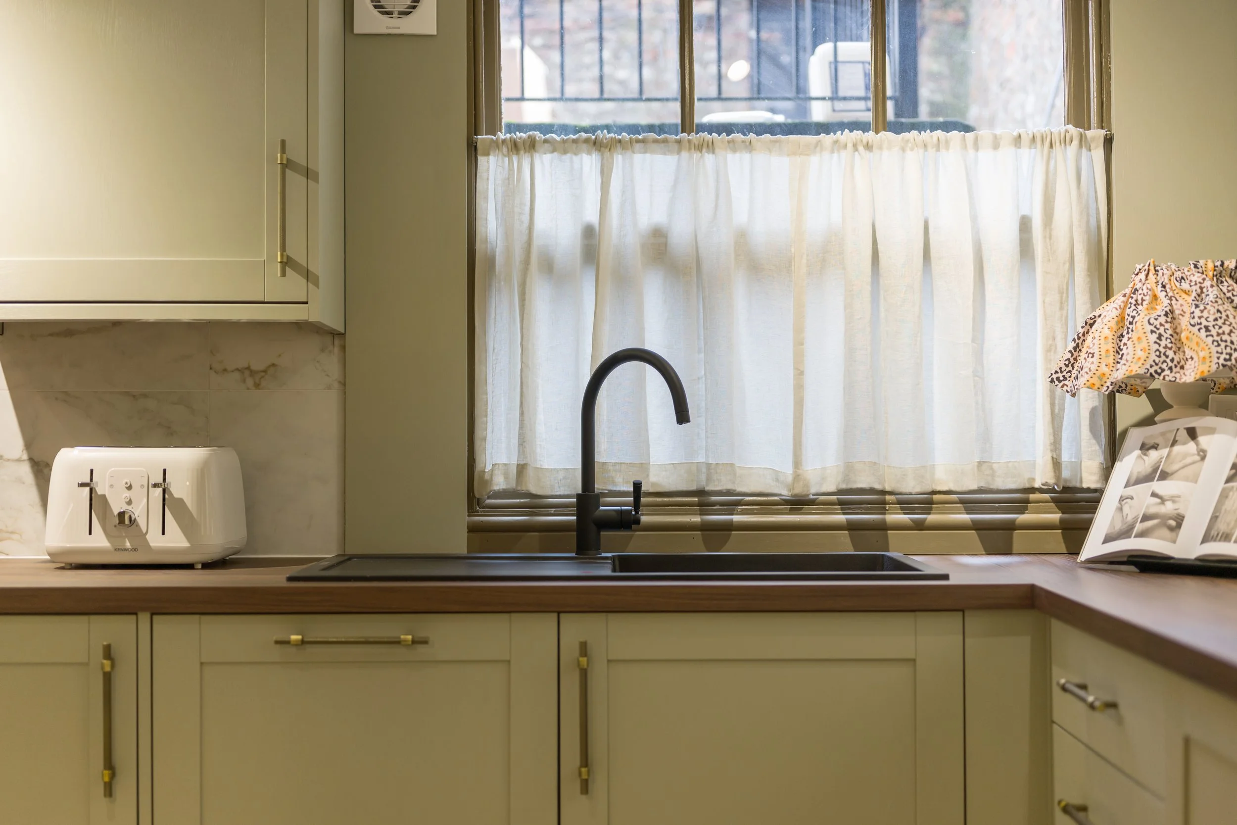 Soft-green heritage kitchen with brass hardware, wooden worktop, black tap and sheer café curtain in a Georgian Quarter renovation by Stiwdio Interior Design.
