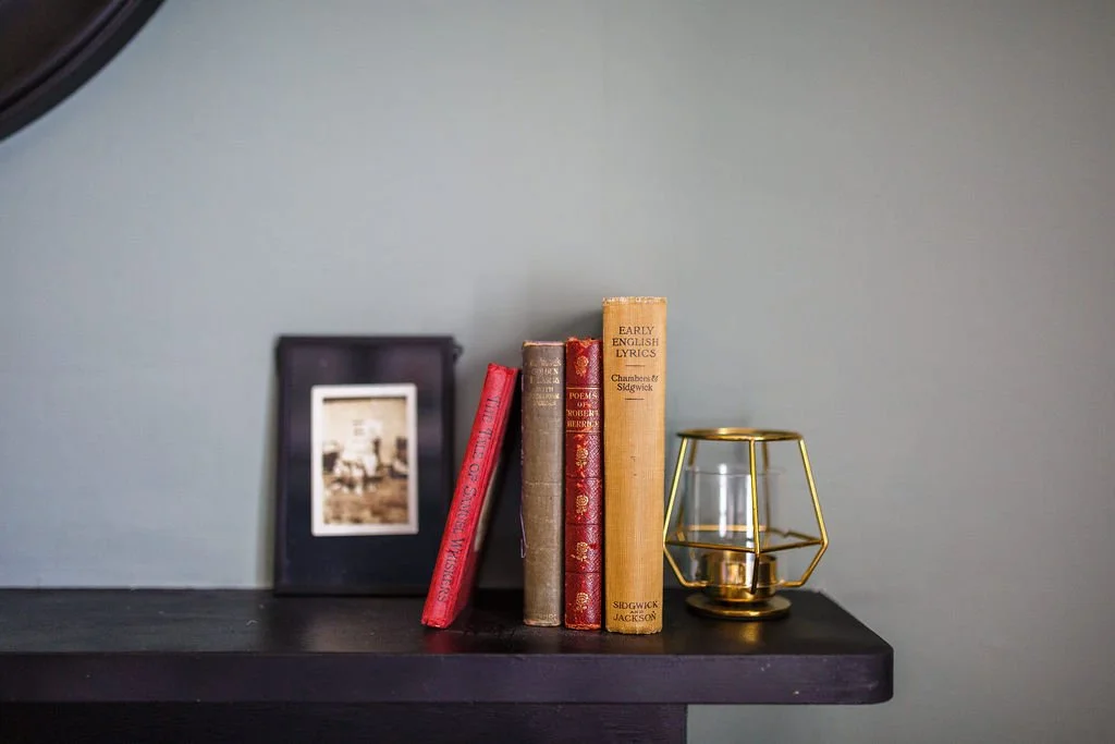 A black shelf holding four old books, a black photo frame with blurred picture, and a gold geometric candle holder with a glass container inside, against a gray wall.