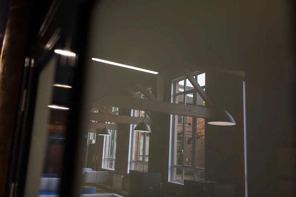 Light-filled heritage office interior within a restored mill building, showcasing exposed timber trusses, industrial lighting and large factory windows—commercial workplace design by Stiwdio Interior Design, North Wales.