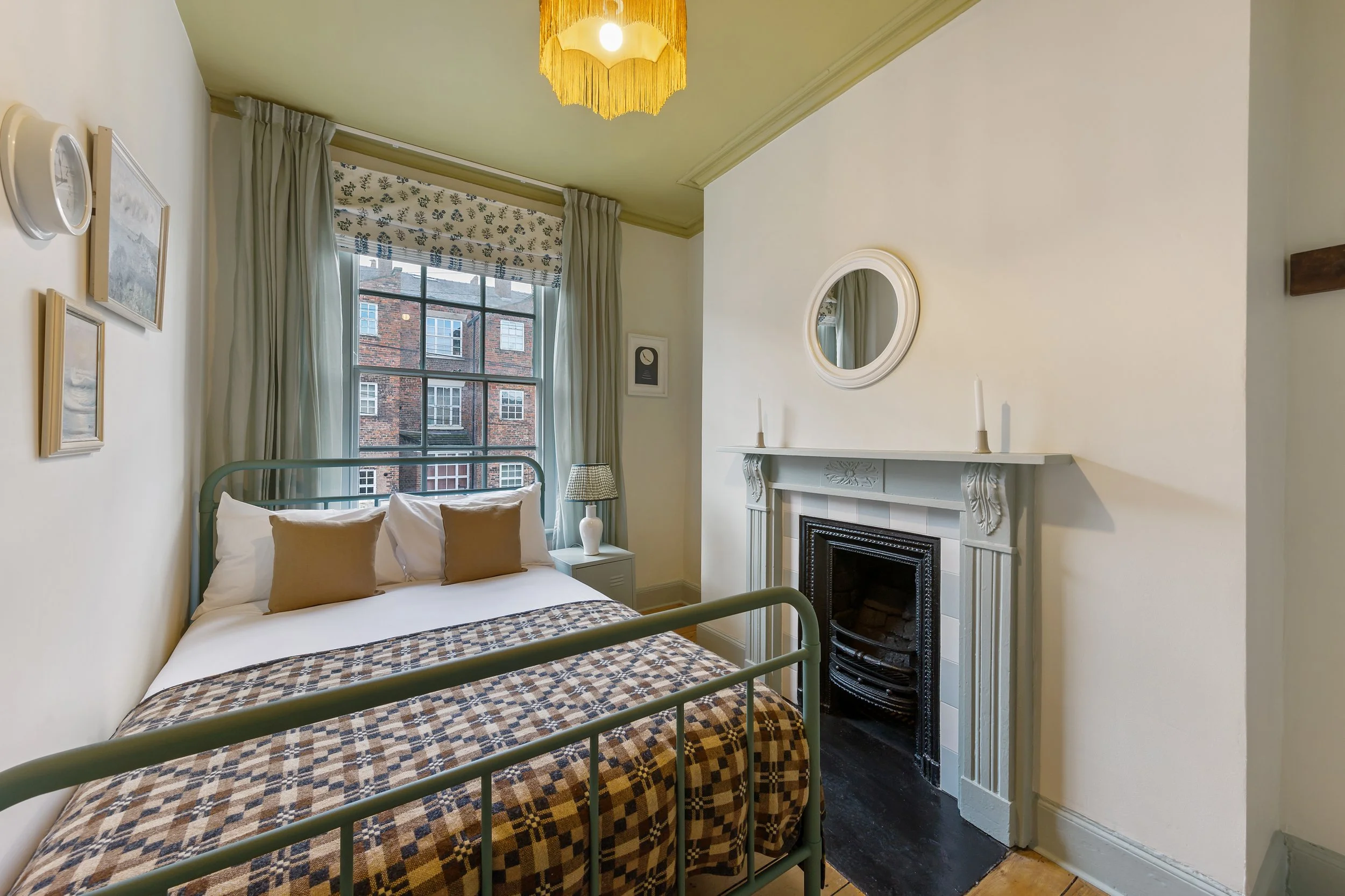 Charming bedroom interior featuring a vintage-style green metal bed frame, patterned throw blanket, and soft neutral bedding. The room includes a traditional fireplace, round wall mirror, pastel décor accents, and large window with curtains, creating
