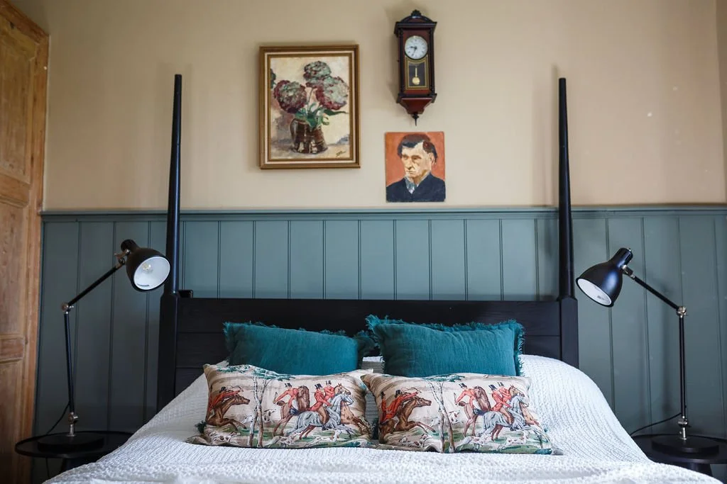 A bedroom with a black four-poster bed, teal pillows, and decorative pillows with horse riding design, wall-mounted lamps, a wall clock, and framed artwork including a floral picture and a portrait.