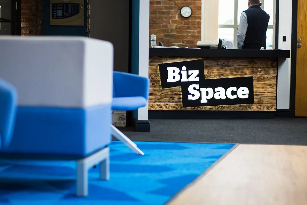 Reception area within a heritage mill office refurbishment for BizSpace, blending exposed brickwork and reclaimed timber with bold contemporary branding. Commercial interior design by Stiwdio, North Wales—creating modern, practical workspaces that ho