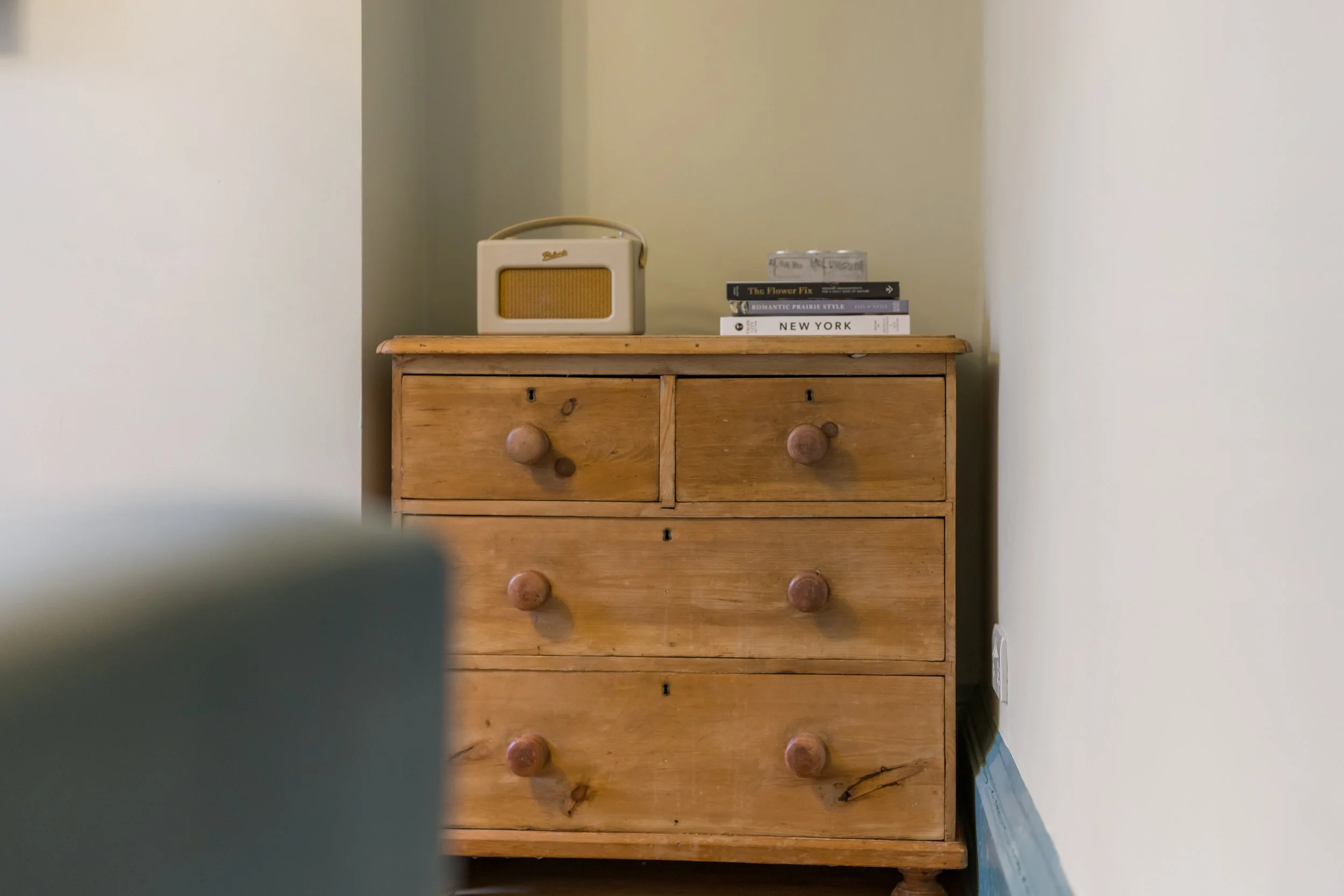 Cozy interior detail featuring a rustic wooden chest of drawers set in a wall alcove, styled with a vintage-style radio and stacked coffee table books. Soft neutral walls and natural wood textures create a warm, minimalist home décor aesthetic with a
