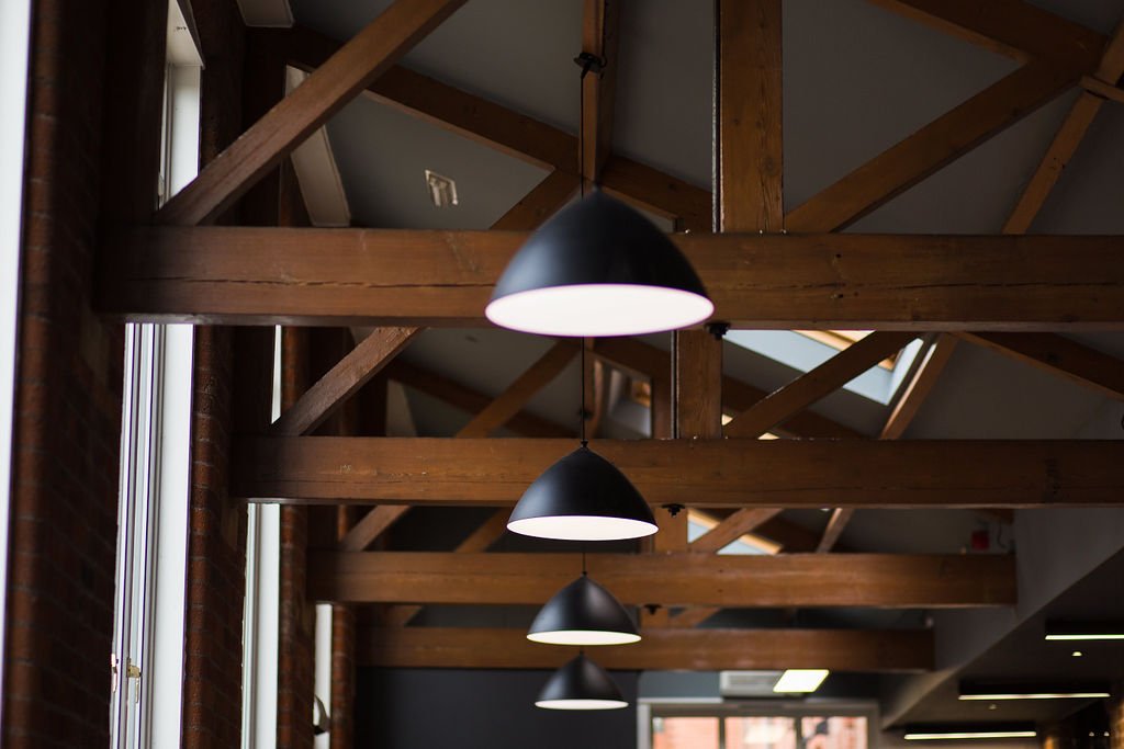 Exposed timber trusses and contemporary pendant lighting in a heritage mill office refurbishment, blending industrial character with modern commercial design by Stiwdio Interior Design, North Wales.