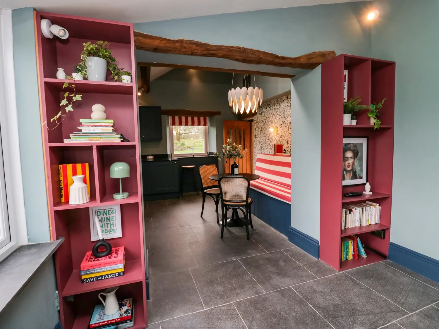 Interior view of a cozy dining area with a round black table and two rattan chairs, decorated with flowers, and built-in booth with red and white striped fabric. The space features pastel blue and green walls, a rustic wooden beam, and pink bookshelves with books and decorative items.