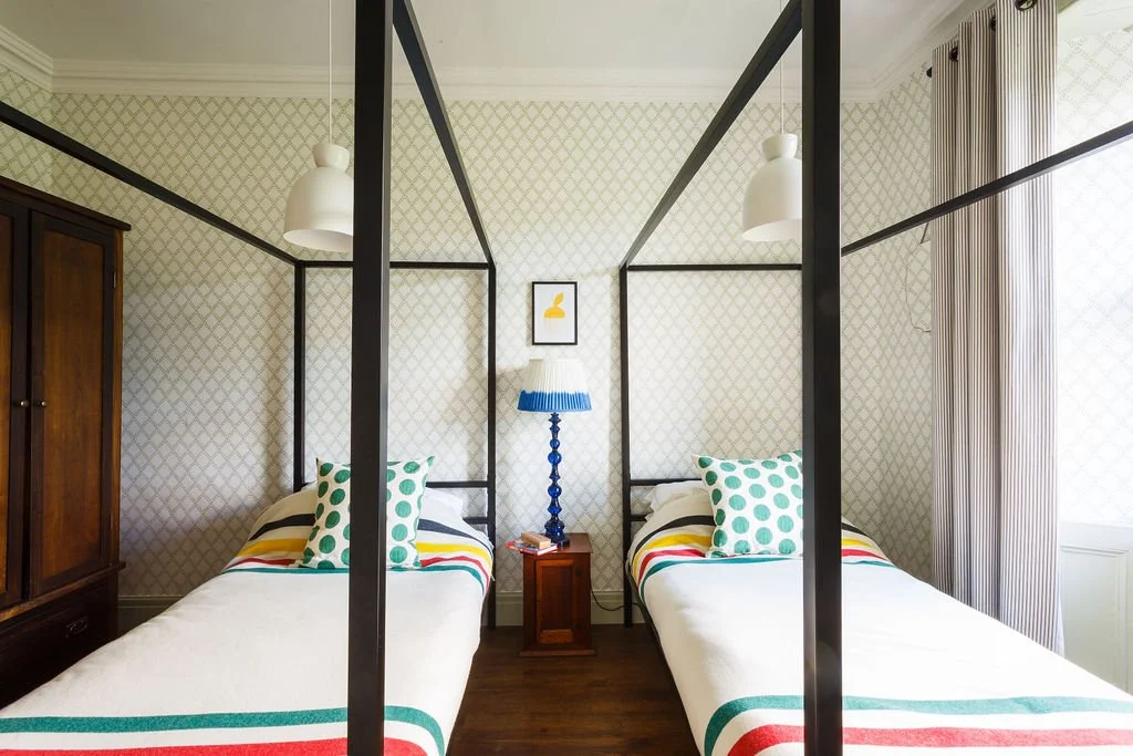 Two twin beds with polka dot pillows in a bedroom with a patterned wall, a wooden wardrobe, a nightstand with a blue lamp, and a framed picture above the headboards.