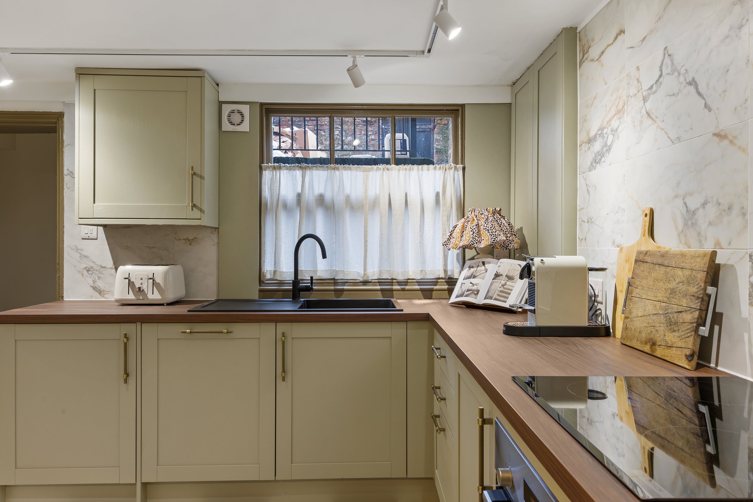 Kitchen with a cafe curtain, sage units and a black tap and marble tiling